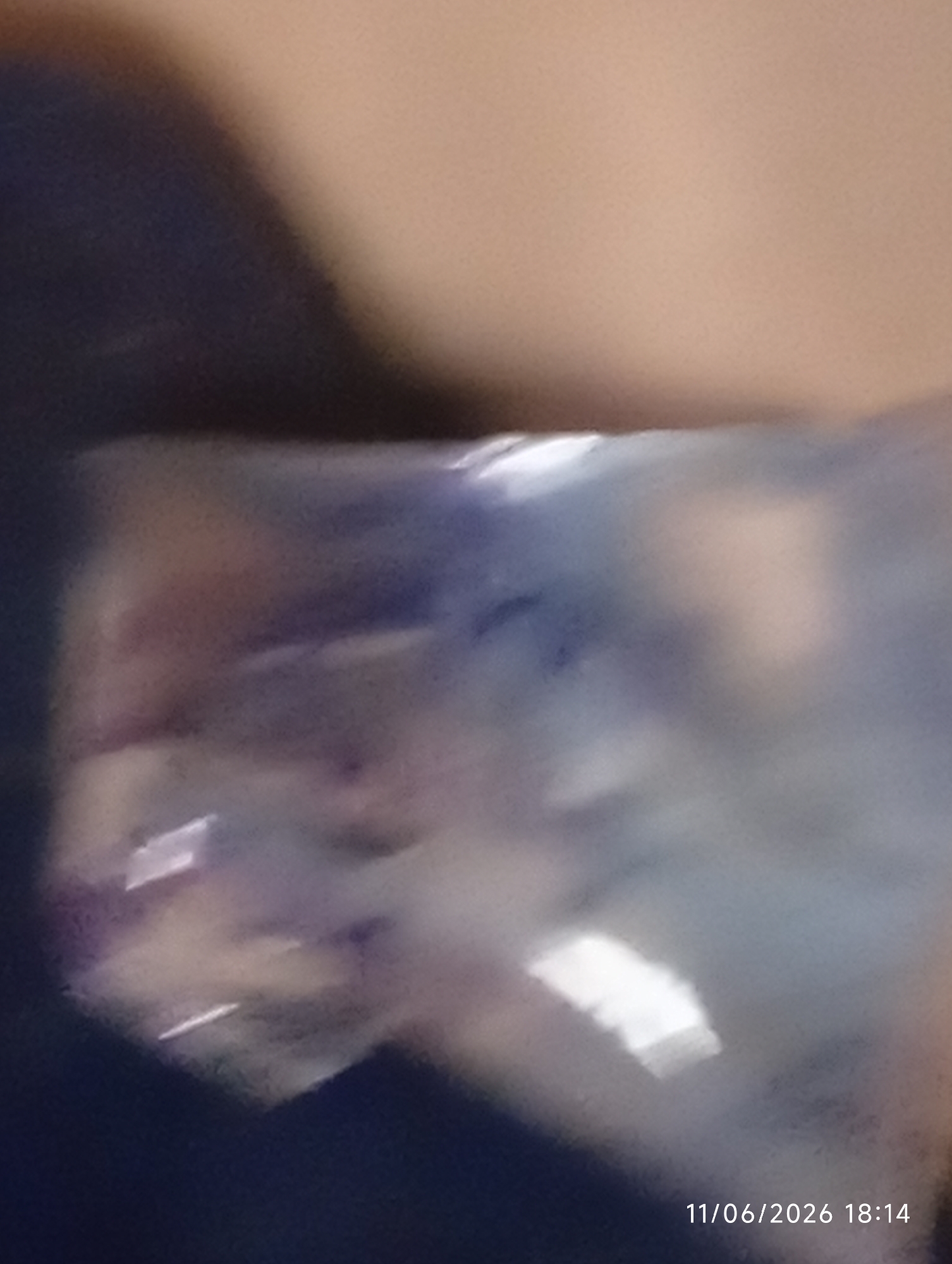 The image shows a blurry, abstract composition of colors and light reflections. The dominant colors are dark blue, beige, and various shades of purple and white, suggesting a reflective or translucent surface, possibly plastic or cellophane. The lighting is uneven, with bright highlights creating abstract shapes. In the bottom right corner, a timestamp reads "11/06/2026 18:14". The image's quality and abstract nature make it impossible to identify specific objects or activities. There is no discernible location in the city of DU, Nigeria, or any identifiable people or actions.