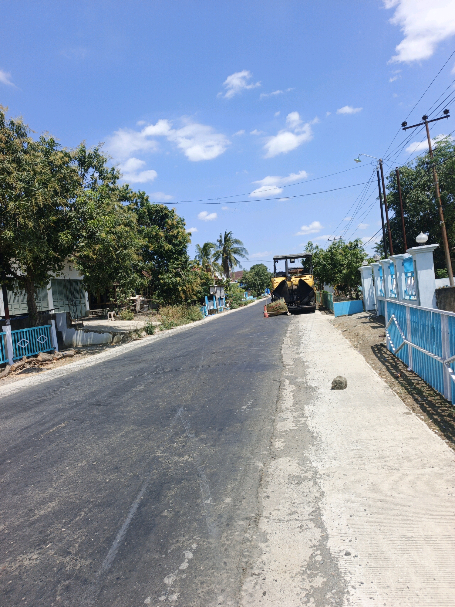 This is a vertical, eye-level, medium shot of a rural road under construction in Talotenreng, Indonesia. The road itself takes up the majority of the foreground, transitioning from dark asphalt on the left to a lighter concrete on the right. The concrete side of the road has visible tire tracks and scattered debris. A single, dark gray rock sits on the concrete shoulder. A bright yellow asphalt paver is positioned on the road, with its rear facing the camera. An orange traffic cone with a yellow warning sign is placed in front of the paver.
To the left of the road, a blue and white fence runs along the edge of a property. Behind the fence, several trees, some with visible foliage and branches, are present. A light-colored building with a green-tinted gate is partially visible behind the trees.
To the right of the road, another section of blue and white fence is visible, leading to a light-colored pillar and wall. Behind this fence, more trees and foliage can be seen, with a light pole and electrical wires extending across the scene. The sky is a bright, clear blue with scattered white clouds. The scene appears to be set during the daytime.
