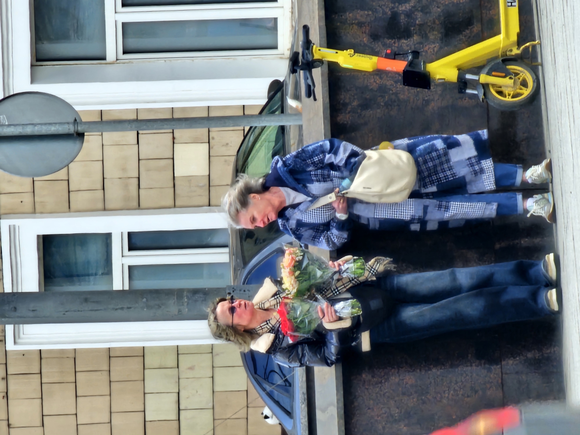 Two women are standing on a sidewalk in Moscow, Russia. One woman is holding flowers. A yellow scooter is parked nearby. A building is in the background.