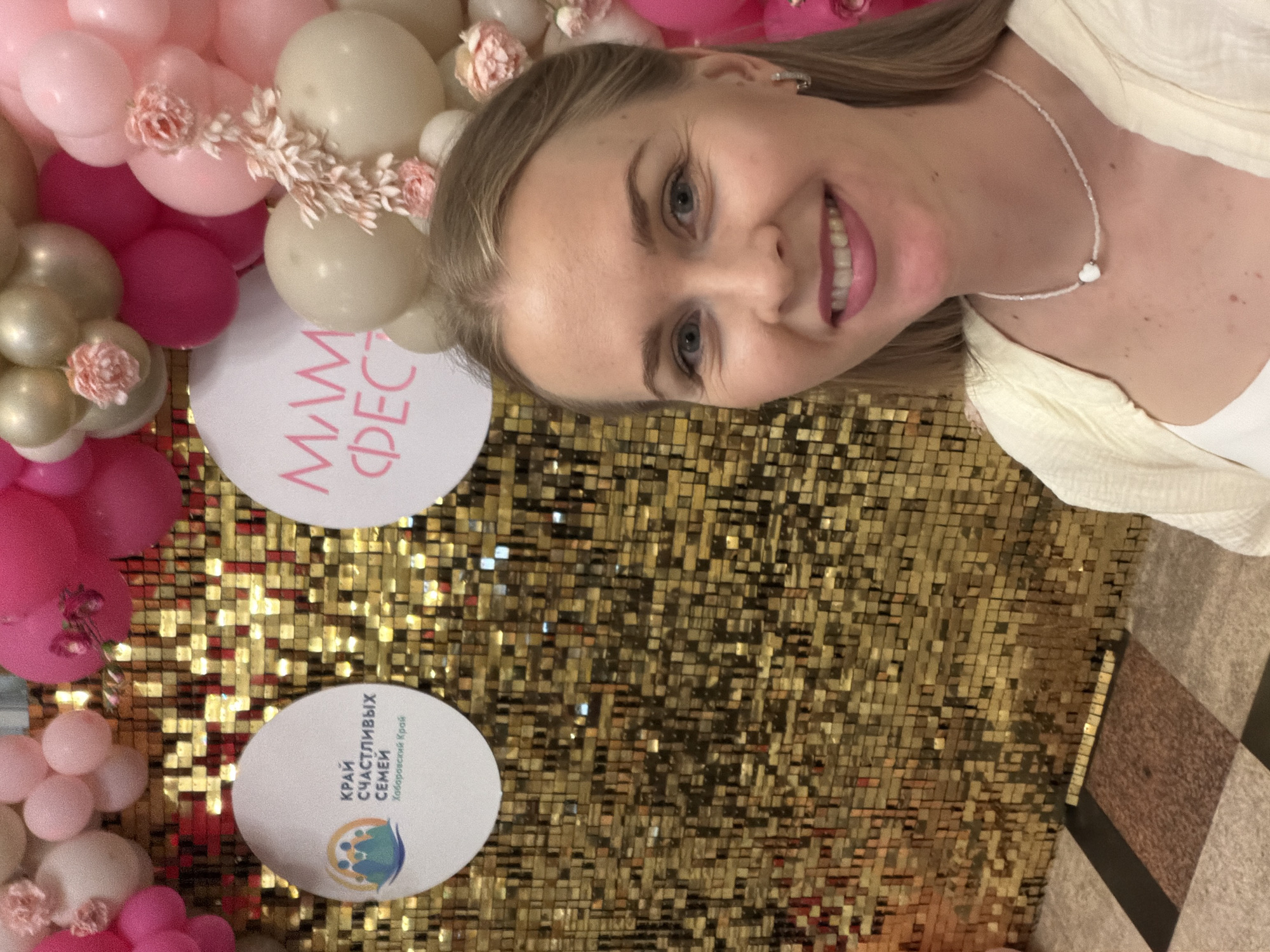 A woman with blonde hair and blue eyes smiles at the camera, taking a selfie in front of a backdrop of gold sequins and pink and beige balloons. She is wearing a white top and a light yellow cardigan, with a beaded necklace featuring a small white heart pendant. To her left, a white circular sign with blue and green graphics and Russian text reads "КРАЙ СЧАСТЛИВЫХ СЕМЕЙ Хабаровский Край," which translates to "Land of Happy Families, Khabarovsk Krai." To her right, another white circular sign with pink text reads "МАМ ФЕСТ," likely indicating a "Mom Fest" event. The overall scene suggests a festive event held in Khabarovsk, Russia, possibly a family-oriented festival.