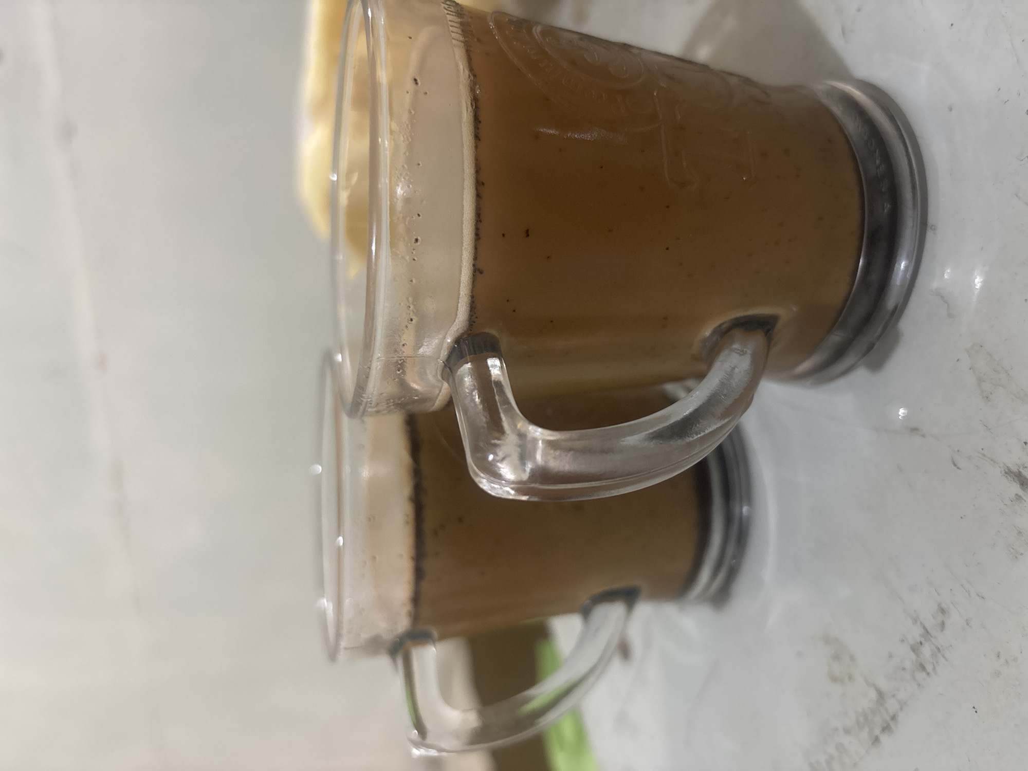 Two glass mugs filled with a light brown beverage are on a white, slightly textured surface. The mugs have clear, curved handles. The beverage in the mugs appears to be a hot drink, possibly coffee or tea, with small bubbles visible on the surface. The background is blurred and appears to be a wall with a lighter texture. No people are visible in the image, and there are no obvious signs indicating the specific location in Krasak, Indonesia. The lighting is soft, suggesting an indoor setting.