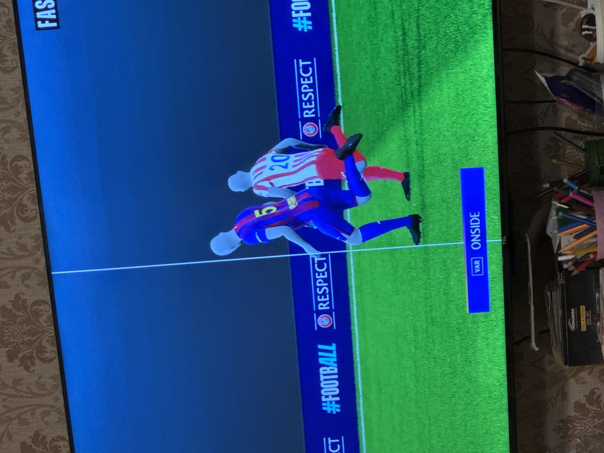 The image depicts a television screen displaying a soccer game. Two players, one in a blue and red uniform with the number 5 and the other in a red and white striped uniform with the number 20, are interacting on a green field. A white line bisects the screen horizontally. The lower portion of the screen displays "RESPECT" and "#FOOTBALL". A blue bar on the right side of the screen shows "VAR ONSIDE". The television is situated in a cluttered environment with visible pencils, a "HOMEBERG" salt and pepper mill set, and other miscellaneous objects. The location is not in Yerevan, Armenia, as there is no information connecting the image to that location.