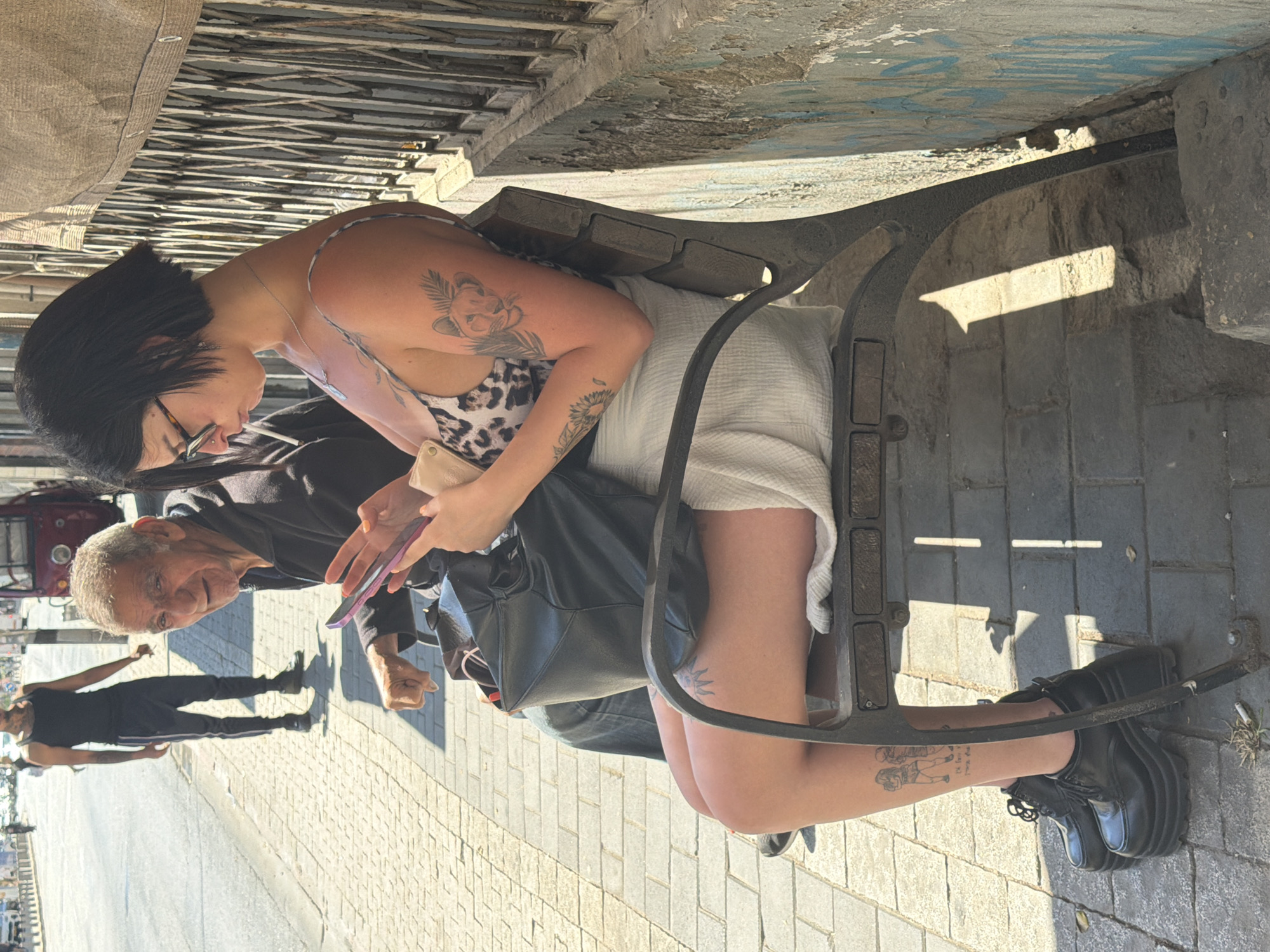 A sunny outdoor scene shows a young woman seated on a metal bench, looking at her cell phone. She has short black hair, wears glasses, a leopard print top, and a white skirt. Tattoos are visible on her arm and lower leg. A black leather purse is next to her. An elderly man stands beside the bench, also looking at the phone. In the background, a man wearing dark clothing walks along the cobblestone sidewalk. The location appears to be a street in Tel-Aviv, Israel.