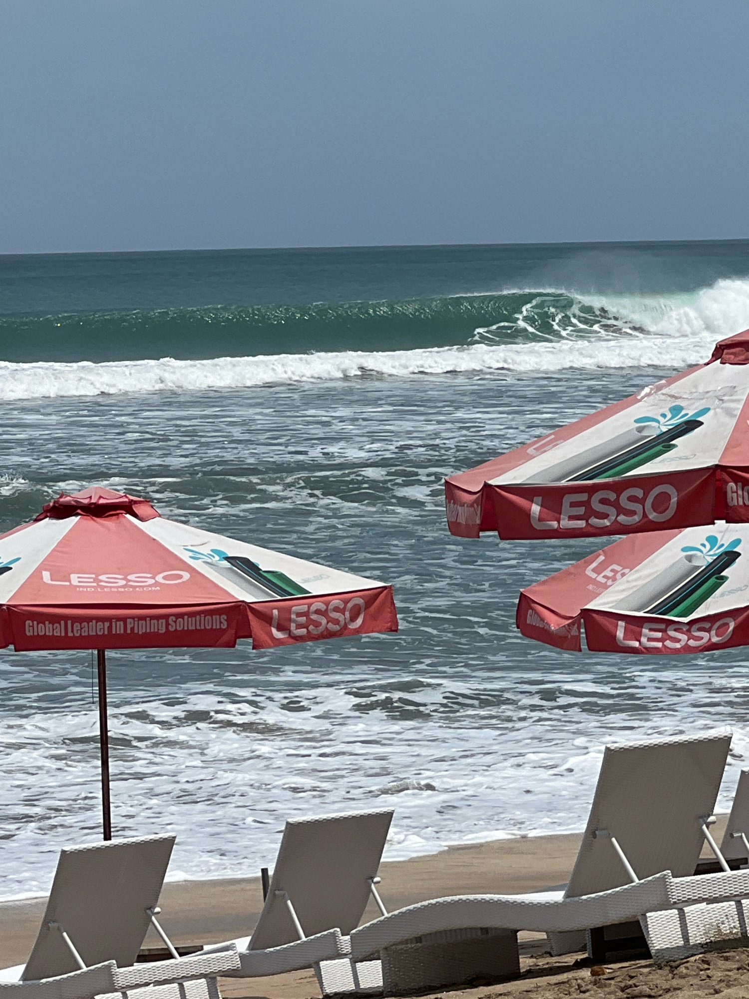 This image captures a beach scene in Kuta, Indonesia. The foreground features several white lounge chairs positioned on a sandy beach. Beyond the chairs, three red and white beach umbrellas are visible, with one prominently displaying the "LESSO Global Leader in Piping Solutions" logo. The umbrellas partially obscure the ocean, where waves are actively breaking. The sea is a deep turquoise color, and the sky above is overcast with a grayish hue. The overall impression is a calm, slightly hazy beach day with commercial branding integrated into the scenery.