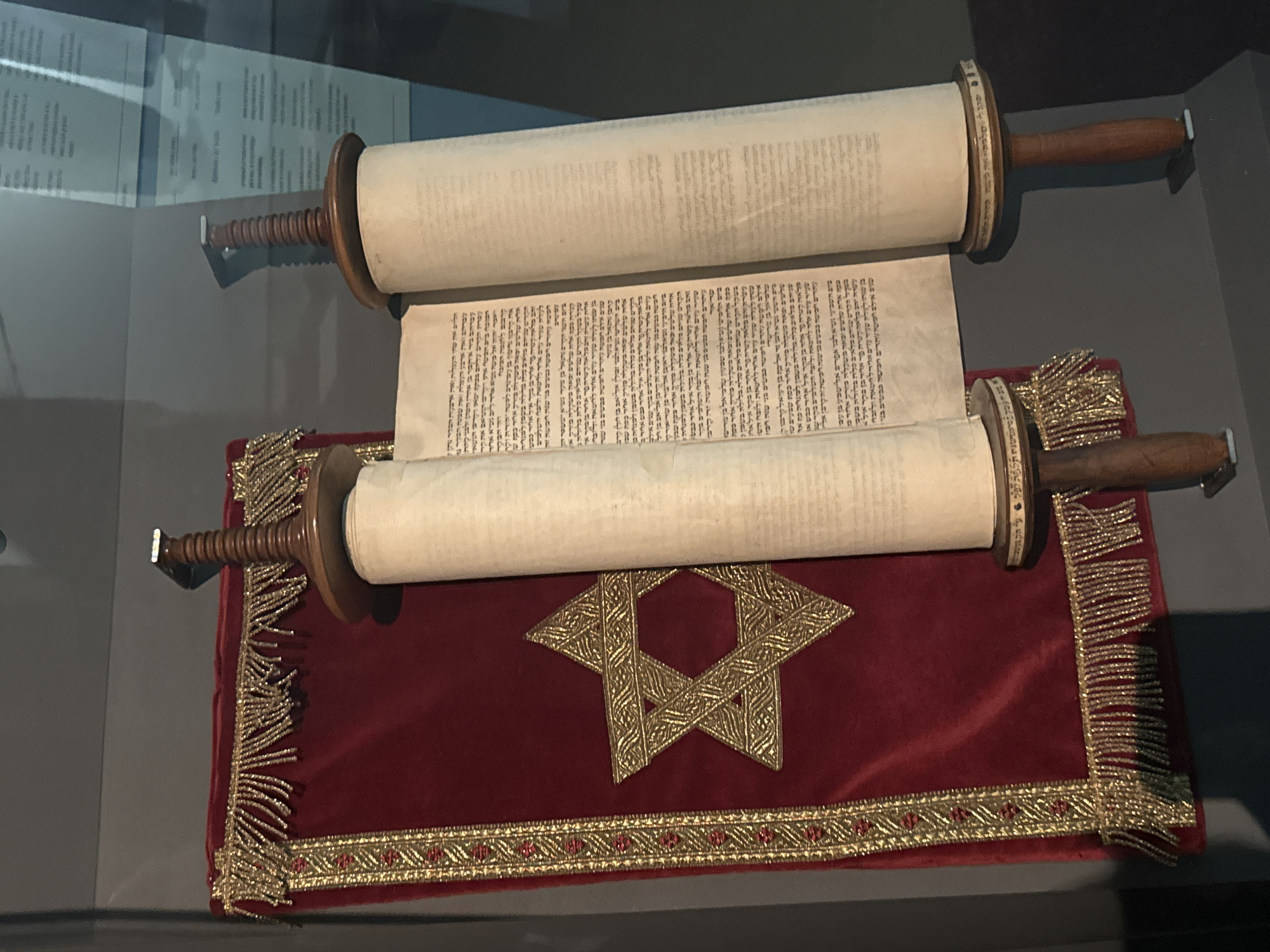 The image depicts a Torah scroll, a sacred text in Judaism, displayed within a museum setting in Bern, Switzerland. The scroll is made of parchment and is unrolled on two wooden rollers, revealing Hebrew script. The Torah is draped over a rich, deep red velvet cloth that is adorned with intricate gold embroidery in the form of a Star of David and decorative borders with fringed tassels. The environment appears to be a dimly lit exhibition space, with a glass partition reflecting some of the surroundings. The focus is on the religious artifact, presented in a manner that emphasizes its historical and cultural significance.