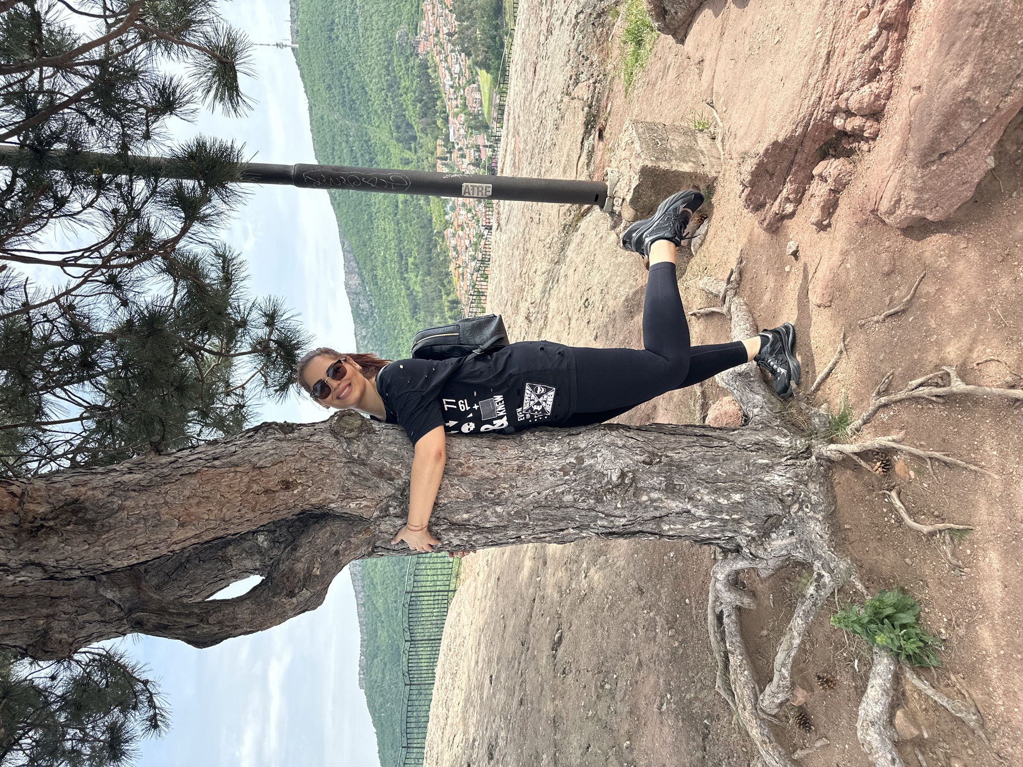 A woman wearing a black t-shirt, black leggings, and black athletic shoes is lying on a tree trunk. The tree is located on a rocky outcrop overlooking a green valley, presumably in the vicinity of Belogradchik, Bulgaria. The tree has exposed roots on the side closest to the ground. The woman is wearing sunglasses and smiling. Her body is oriented horizontally, with her head near the tree branches on the left and her feet near the rocky edge on the right. A metal pole or railing runs above her feet. In the background, there are trees and a town.