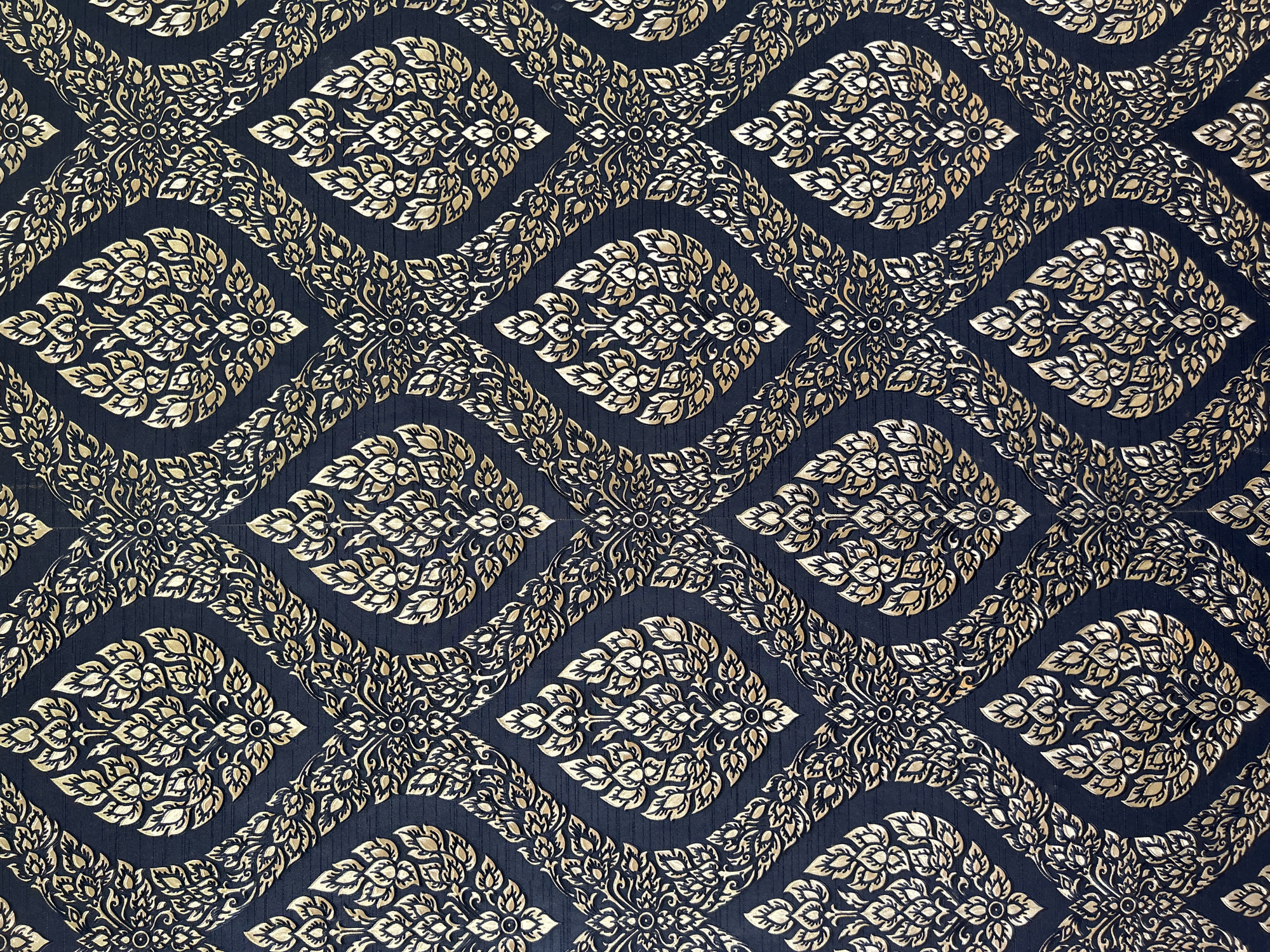 The image displays a detailed view of a dark blue fabric with an intricate, embossed golden pattern. The pattern is a repeating design featuring teardrop-shaped motifs filled with stylized floral elements and leaves. These motifs are arranged in a diamond grid, connected by narrow, ornate bands that also feature leaf-like designs. The golden pattern has a metallic sheen, highlighting its raised texture against the dark background. The image is a full frame, top-down shot of the fabric, with no visible extraneous elements or background context suggesting a specific location within Thai Ko Subdistrict, Thailand, or any other identifiable setting.