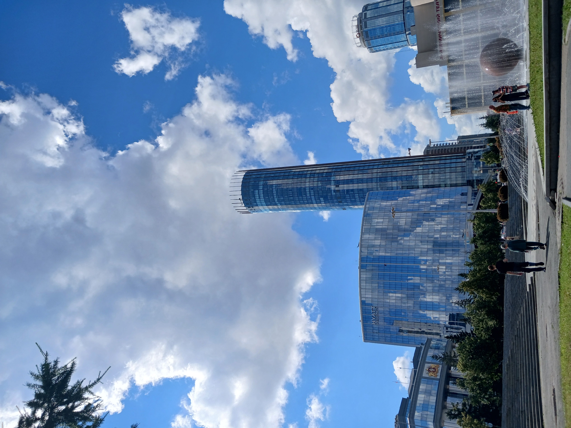 This is a photograph of a modern city scene in Yekaterinburg, Russia. The image is oriented vertically, showing a view looking up towards tall, glass-facade buildings against a bright blue sky with fluffy white clouds.
In the foreground on the right, there is a paved walkway and a grassy area. People are visible walking along the walkway, some near a water feature that includes a large spherical object. The water feature is producing a spray, creating a dynamic element in the scene. The buildings in the background are contemporary, featuring reflective glass that mirrors the sky and clouds. One building has the text "HYATT REGENCY" visible on its facade. Another building appears to be an older structure with ornate details above its glass facade.
A lone pine tree is visible in the lower left corner, reaching upwards. The lighting suggests it is daytime, likely mid-morning or afternoon, with bright sunlight casting shadows. The weather appears to be clear and pleasant, with a good mix of sun and clouds. The overall mood is one of urban vitality and modern architectural presence.