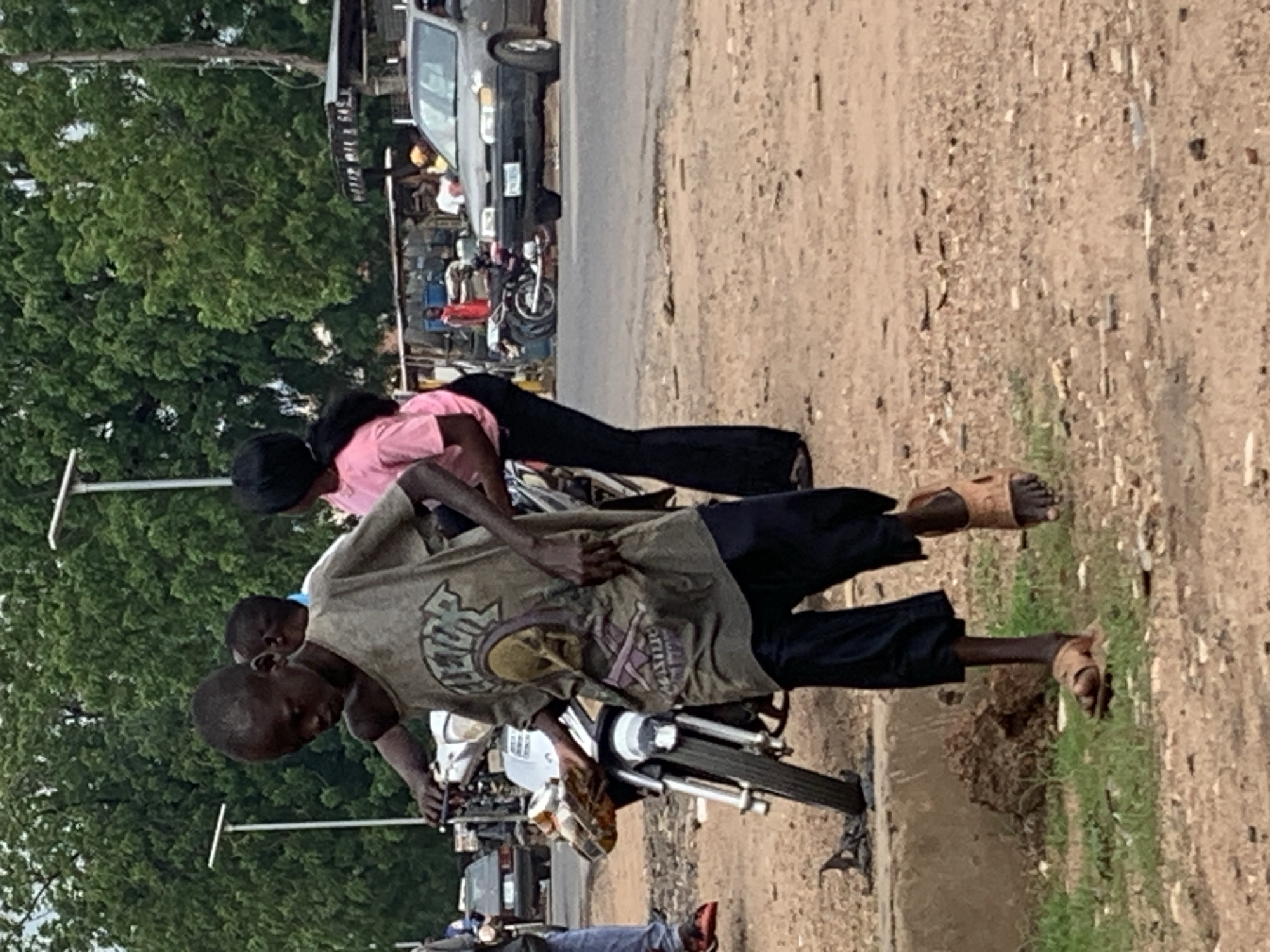 This image captures an outdoor street scene in Shendam, Nigeria, under daylight conditions. The foreground consists of reddish-brown dirt and gravel with sparse patches of green grass, leading to a paved asphalt road in the mid-ground. Dense green foliage from large trees forms the background.
The central figure is a dark-skinned male child or young adolescent with a thin build and short hair. He wears a faded, dirty, light brown t-shirt featuring a dark graphic print (possibly a skull or abstract design), dark blue or black baggy trousers, and light brown open-toed sandals. He is positioned slightly off-center to the left, holding the handlebars of a silver-colored motorcycle with his left hand and a clear plastic container with yellow contents in his right hand. He appears to be walking forward or standing beside the motorcycle.
Behind the central figure, partially obscured, is another dark-skinned individual wearing a light-colored top. To the right of the central figure, a dark-skinned female wearing a pink t-shirt and dark, flared pants is partially visible, oriented away from the viewer.
On the paved road in the mid-ground right, a dark gray sedan (appearing to be a Honda Accord) is present, with its front facing the viewer's left. A white license plate with partially visible characters is affixed to its front bumper. Behind this car, a person in a white t-shirt is visible. Further left, on the same paved road, several motorcycles are parked, and a person in a red top stands near them. Behind these parked motorcycles and people, a makeshift structure with corrugated metal roofing is visible, bearing partial text "HERER OIL & GAS".
In the background, two tall, slender white streetlights with horizontal fixtures stand above the tree line. A utility pole is visible on the far right edge of the frame. The overall impression is a bustling, informal street environment.
