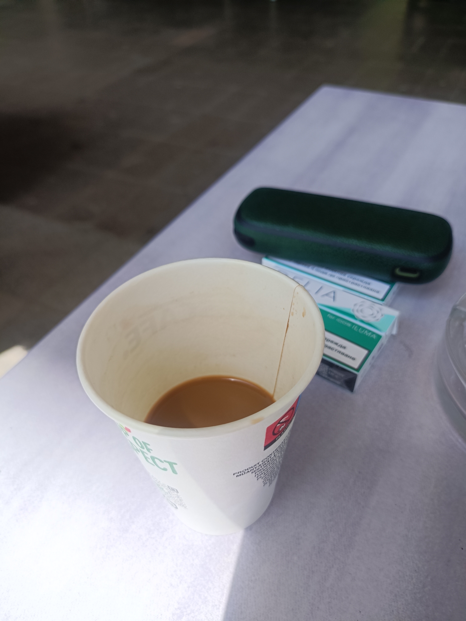 The image presents a close-up, slightly elevated view of various items arranged on a light-colored, textured surface, possibly a tablecloth. In the foreground, a white paper cup contains a small quantity of light brown liquid, likely coffee or tea. The cup exhibits a vertical crease or crack on its rim and features partially visible text, including "OF FECT" and a QR code.
Behind the cup, positioned to the right, is a dark green elongated protective case. It rests upon two light green and teal-colored boxes, stacked vertically. These boxes are clearly labeled "TEREA for IQOS ILUMA," indicating they are for heated tobacco products. Visible text on the packaging includes "ILUMA," "TEREA for IQOS ILUMA," along with Cyrillic words such as "ПРОДАЖА" (sale) and "ОГРАНИЧЕНА" (limited), which are common on tobacco product warnings in regions like Plovdiv, Bulgaria. A portion of a clear plastic container, possibly an ashtray or a lid, is visible on the far right.
The background is softly out of focus, showing a darker, undefined surface. The lighting suggests daylight, with subtle shadows cast by the objects, indicating an indoor or sheltered outdoor setting. No people are visible, and the scene depicts a quiet, personal moment, possibly a break for coffee and a heated tobacco product.