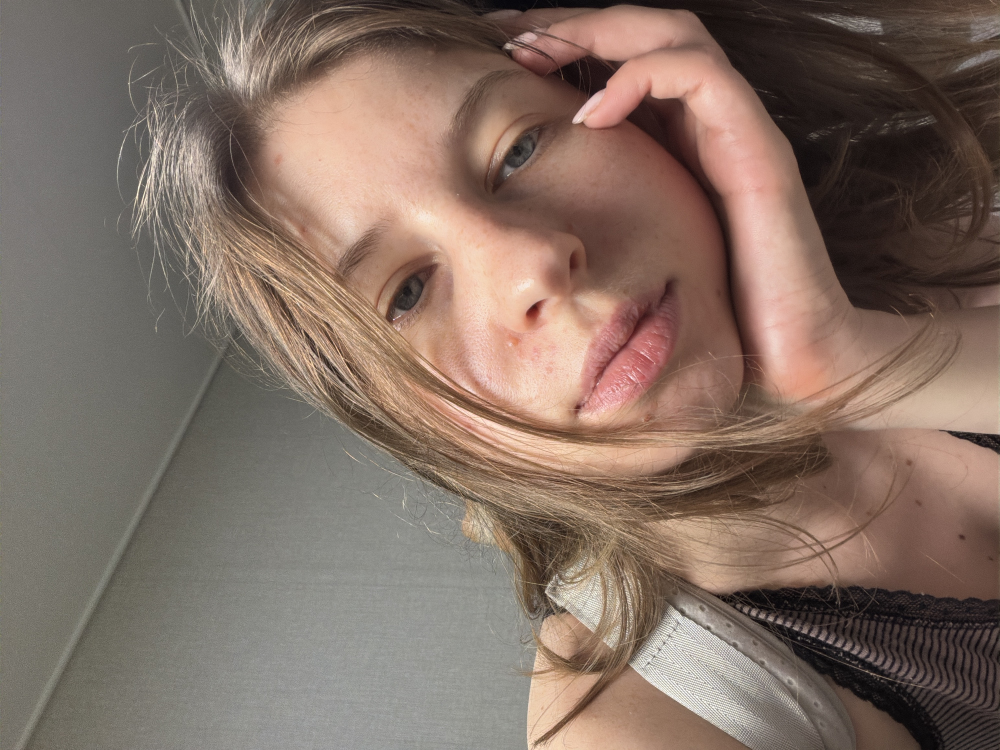 A young woman with long, light brown hair is shown in a close-up, angled selfie. Her hand is resting on her face, near her temple, with her fingers gently touching her cheek. Her eyes are open and looking towards the camera, and her lips are slightly parted. She has light skin with freckles and a few blemishes.
The background appears to be an indoor setting with a textured, gray wall. A portion of what seems to be clothing, possibly a bra with lace trim and straps, is visible on the right side of the image. The lighting suggests it could be daytime, possibly with natural light coming from the side, casting soft shadows. The overall mood is relaxed and intimate, with a hint of vulnerability conveyed by the subject's pose and direct gaze. No text is visible in the image.