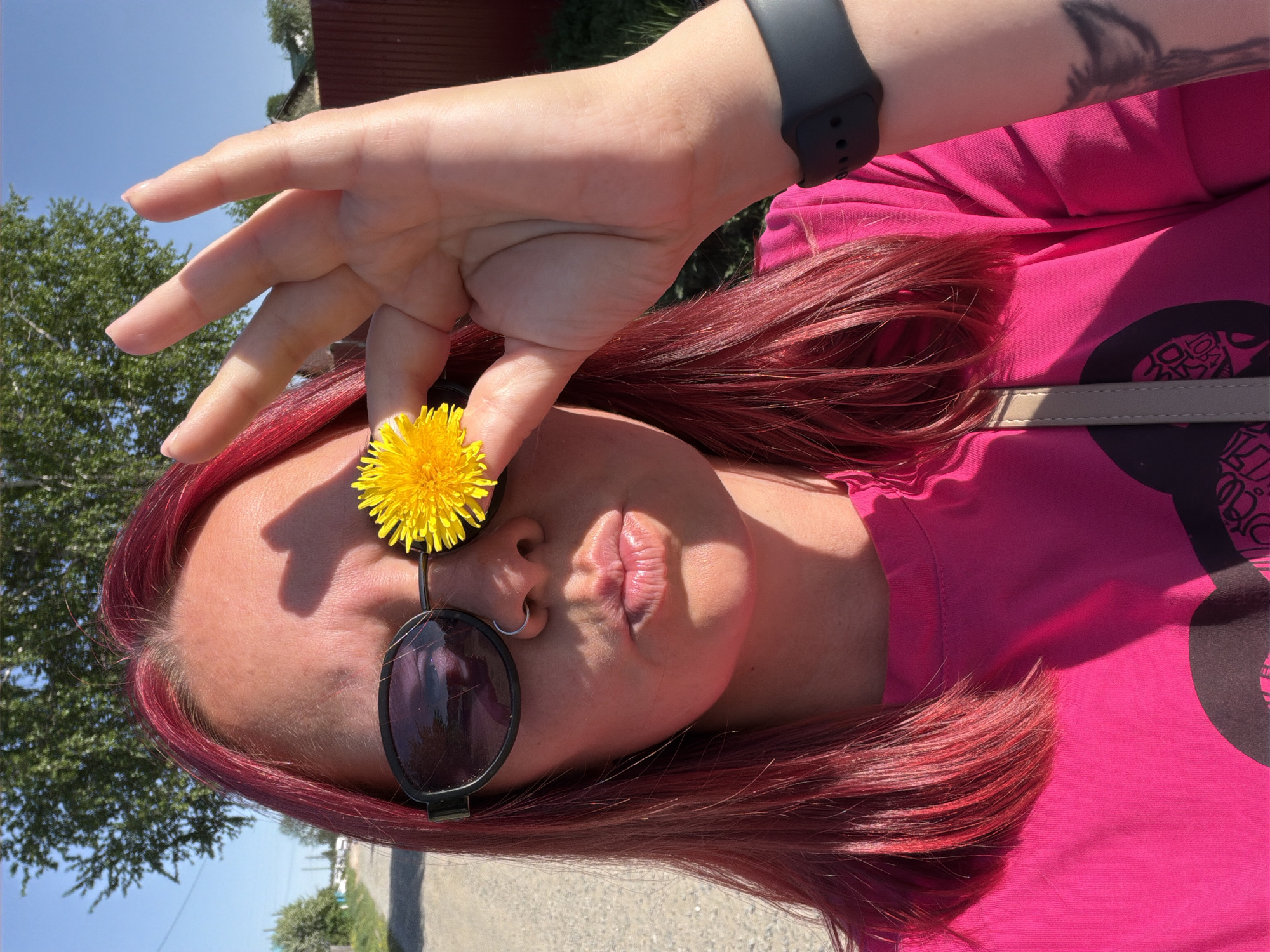 A person with red hair is holding a dandelion in front of their eye. They are wearing sunglasses and a pink shirt. The location is Chaikovsky, Russia. There is a street and some trees in the background. The person is making a duck face. They are wearing a black smartwatch on their left wrist and have a tattoo on their left arm.