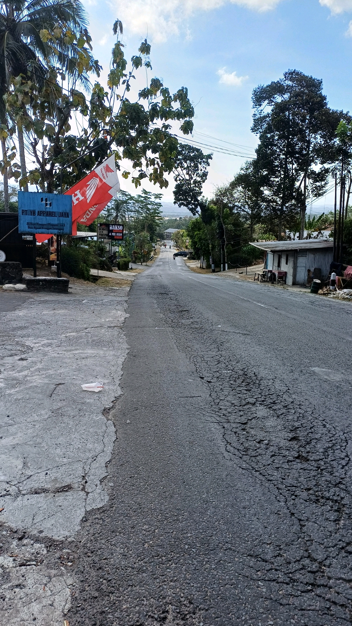 The image shows a street scene in Tengahan, Indonesia. A paved road stretches into the distance, with cracks and imperfections visible on its surface. On the left side of the road, a concrete sidewalk meets the asphalt. A blue sign with white lettering reads "PT. YB APPAREL JAYA." Next to it, a red and white flag with a logo is attached to a pole. Further down the road, there are several small buildings, trees, and power lines. A few people are visible in the distance near one of the buildings. A car is parked on the side of the road. The sky is blue with some scattered clouds.