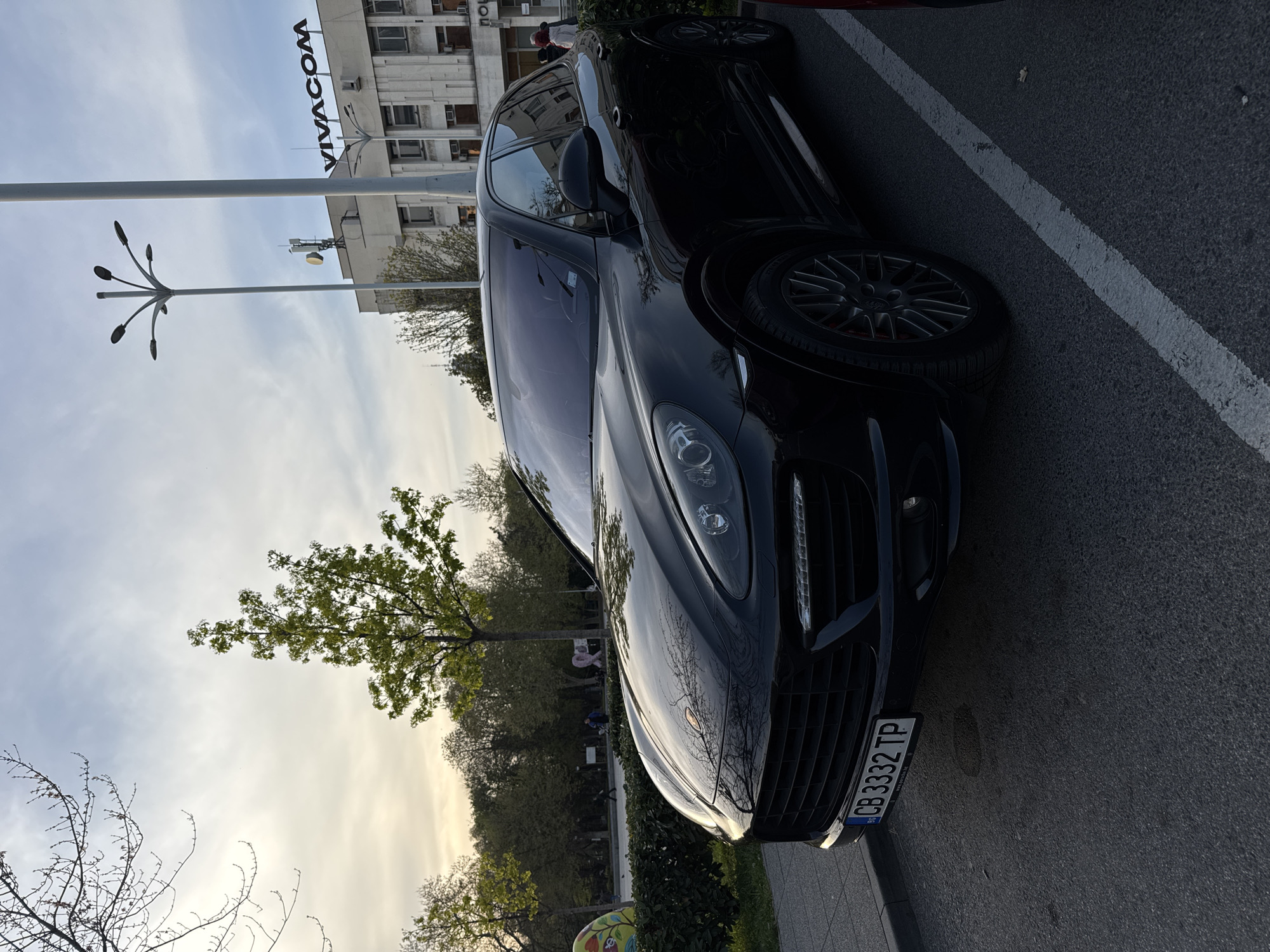 The image depicts a black Porsche Cayenne parked on the side of a street in Plovdiv, Bulgaria. The car is positioned at an angle in a parking space delineated by a white line on the asphalt. The license plate, partially visible, reads "CB 3332 TP".
In the background, there's a building with the "VIVACOM" sign on top, indicating the presence of a telecommunications company. A stylized street lamp stands nearby. A tree with budding green leaves is visible. A public space with a few figures is also visible. The sky is pale with some cloud cover. The image appears to have been taken during daylight hours, possibly in the late afternoon or early evening, judging by the lighting. The scene is urban, blending elements of commercial and public space.