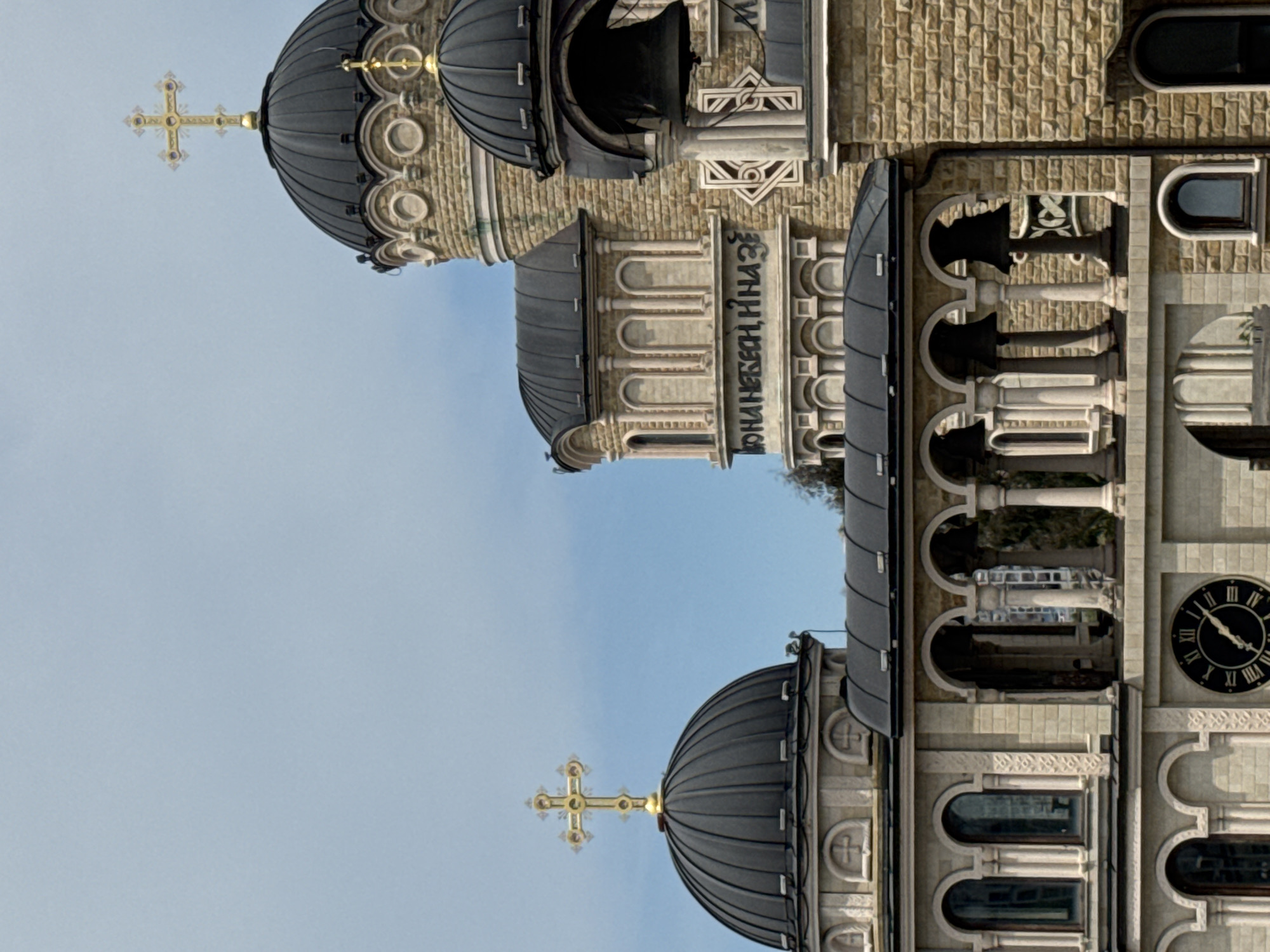 The image depicts the exterior of the Cathedral of Michael the Archangel located in Sochi, Russia. The architecture is characterized by its domed roofs, decorative brickwork, arched windows, and columns. Prominent features include golden crosses atop the domes and a clock on one of the building's towers. The building is situated against a light blue sky.