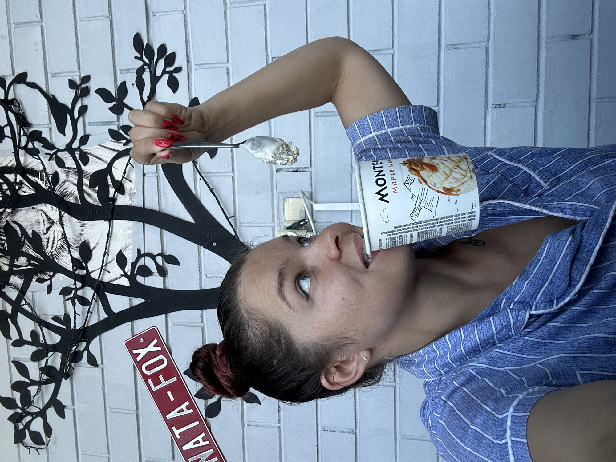 The media file shows a young woman in a blue and white striped pajama top, with her hair tied up in a bun. She is holding a spoon with ice cream in her right hand and is about to eat it from a carton of "MONTE MAPLE WALNUT" ice cream. The woman has her eyes looking up and to the side, with a playful expression. Behind her, a black cut-out tree with branches and leaves is visible against a white brick wall. A red sign with white text that reads "NATA-FOX-" is also present on the left side of the image. The lighting suggests it might be daytime, possibly outdoors given the textured wall that resembles an exterior brick surface. The overall scene appears casual and relaxed, with the woman enjoying a treat. There is no discernible weather information.