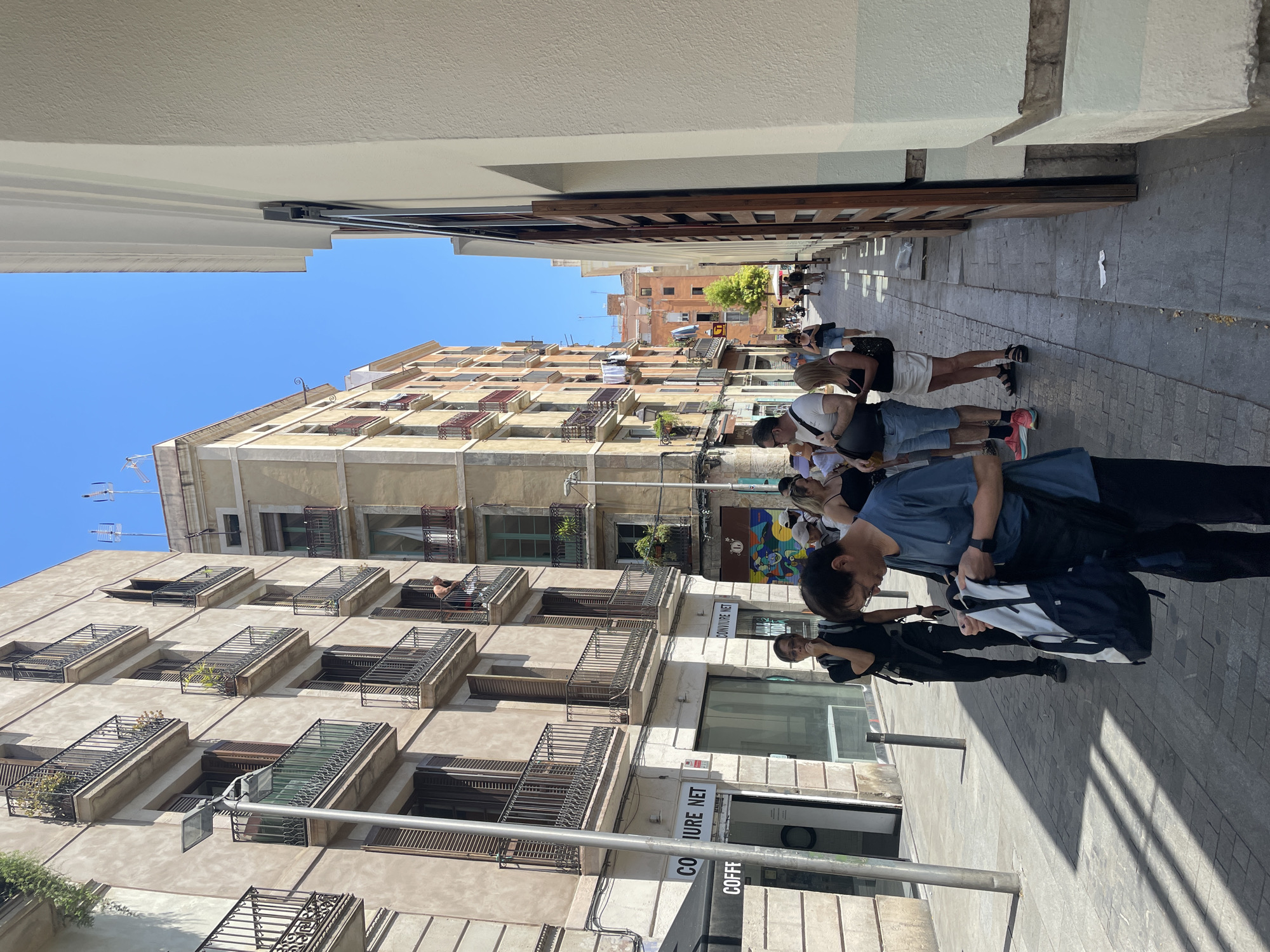 This image captures a sun-drenched street scene in Barcelona, Spain, likely in the Gothic Quarter or El Born, characterized by its narrow cobblestone alleyways and historic architecture. A group of tourists, dressed in casual summer attire, are visible on the right side of the frame, appearing to be a guided tour group pausing or gathering.
In the foreground, a man in a blue t-shirt is looking down at a backpack he is holding. To his left, another man wearing a black t-shirt and backpack stands with his hand near his mouth. Further back, a woman in a white skirt and black top is talking to a man in a blue shirt and shorts, and other individuals are dispersed along the street.
The buildings lining the street are typical of Barcelona's urban landscape, with multi-story structures featuring balconies with wrought-iron railings, wooden shutters, and cream-colored facades. Laundry is seen hanging from some of the balconies, a common sight in older European cities. The street itself is paved with gray cobblestones, and shadows are cast by the buildings and the people.
On the left side of the image, a storefront displays signage for "COIFFURE NET" and "COFF". The overall atmosphere suggests a warm, bright day with a moderate number of people present in a lively urban setting.