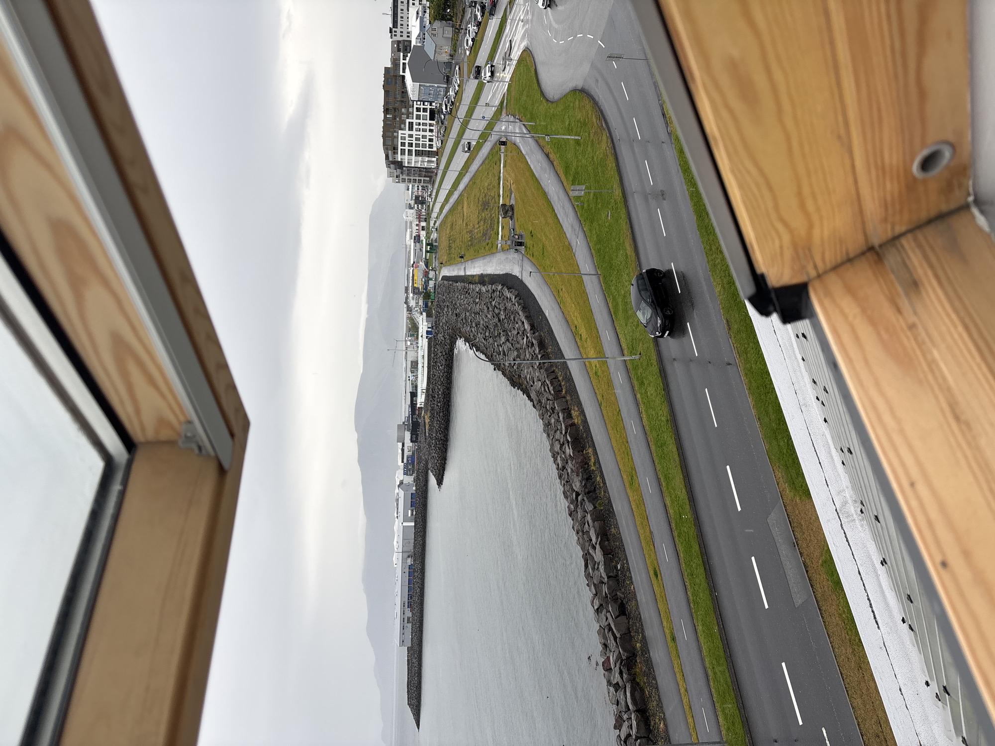 This image provides an overhead view of a coastal road and harbor area in Reykjavik, Iceland, as seen through a skylight. The foreground features the wooden frame of the skylight, partially obscuring the scene below. The main focus of the image is a curved road with multiple lanes, bordered by green grassy areas and a paved pedestrian path. A dark-colored hatchback car is visible driving on the road, its headlights on. To the left of the road, a rocky breakwater separates the water from the road and path. Beyond the breakwater, a body of calm, gray water is visible, with industrial buildings and a distant mountain range forming the backdrop. The sky is overcast and appears to be raining lightly, as evidenced by the wetness of the road and the presence of water droplets on the skylight. The architecture of the buildings in the distance suggests an urban environment, likely part of Reykjavik's industrial or commercial district.