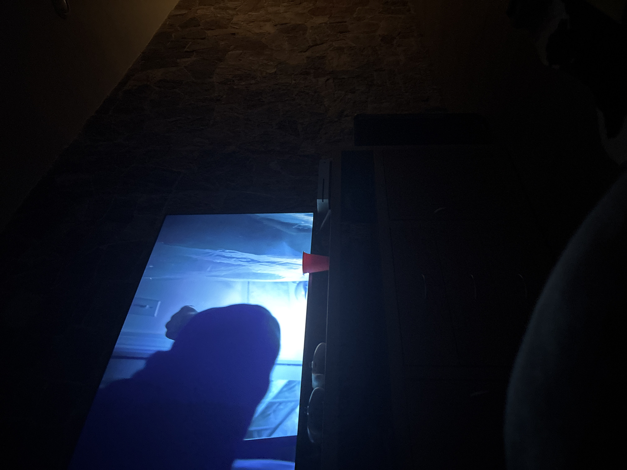 The scene is dimly lit, likely indoors. The primary focus is a glowing screen, possibly a television or computer monitor, which is displaying an image of what appears to be a person, heavily shaded in blue. A portion of the screen is obscured by the edge of a dark, wooden cabinet or piece of furniture. A small, red, cylindrical object is visible near the top edge of the screen and cabinet. The floor and wall are constructed with a stone or rock material.
