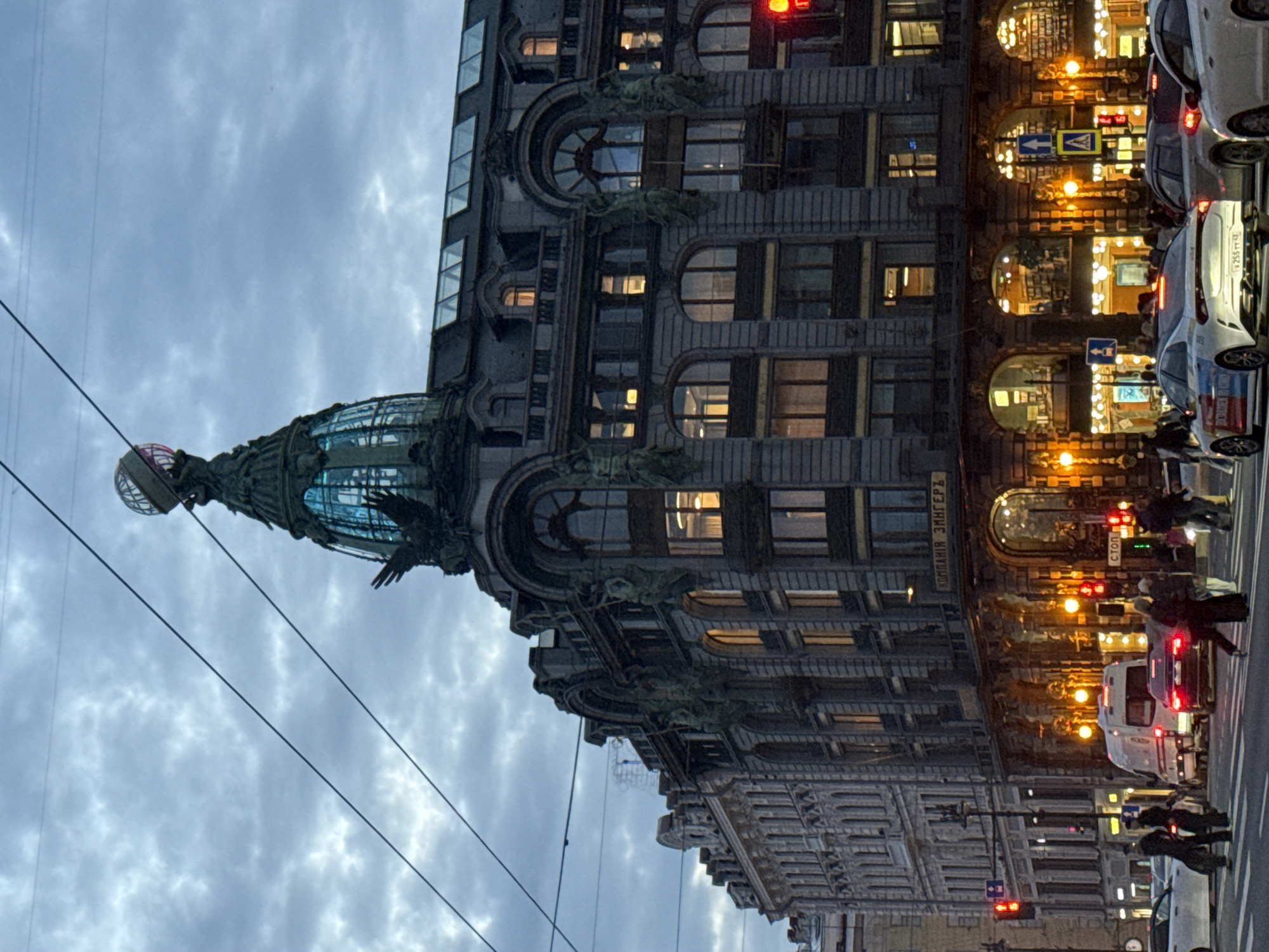 The image shows the Singer House, also known as the Dom Knigi, in Saint Petersburg, Russia, in the evening. The building's distinctive globe-topped dome and ornate facade are visible against a cloudy sky. The street in front of the building is busy with traffic, including cars and a white van. Pedestrians are crossing the street at a crosswalk. Streetlights and illuminated windows cast a warm glow on the scene. The overall impression is a bustling urban environment at dusk.