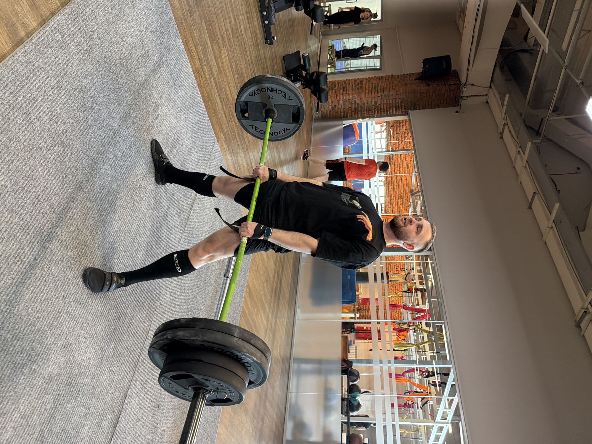 A medium shot depicts a man performing a Zercher squat with a barbell in a gym setting. He is wearing black athletic clothing, including shoes, socks, shorts, and a t-shirt, along with wrist wraps. The barbell, which has green padding around the bar, is loaded with multiple black weight plates on each side. The man is looking upward as he executes the squat.
The gym features a carpeted floor, with other gym equipment visible in the background. In the distance, people can be seen working out. One wall is brick and windows run along the other wall. The location may be in the city of Chaikovsky, Russia, but that cannot be determined from the image alone.