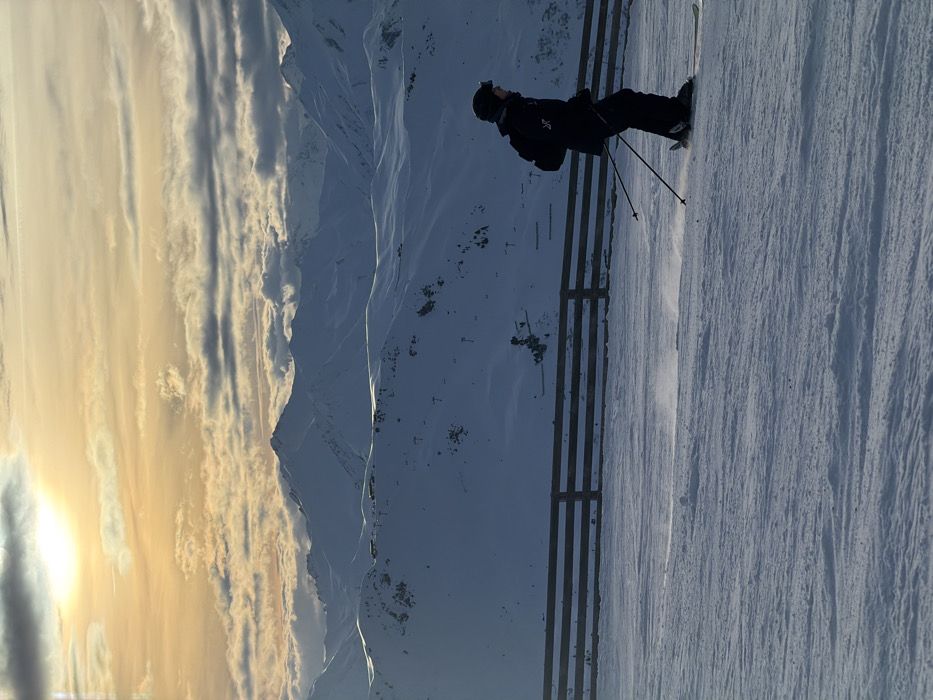 A skier descends a snowy slope alongside a safety fence. Distant mountains are visible behind the fence, nestled amongst a sky filled with dramatic clouds. A bright glow illuminates the left side of the frame, suggesting a sunrise or sunset. The scene portrays a winter landscape with an active skier navigating the terrain.