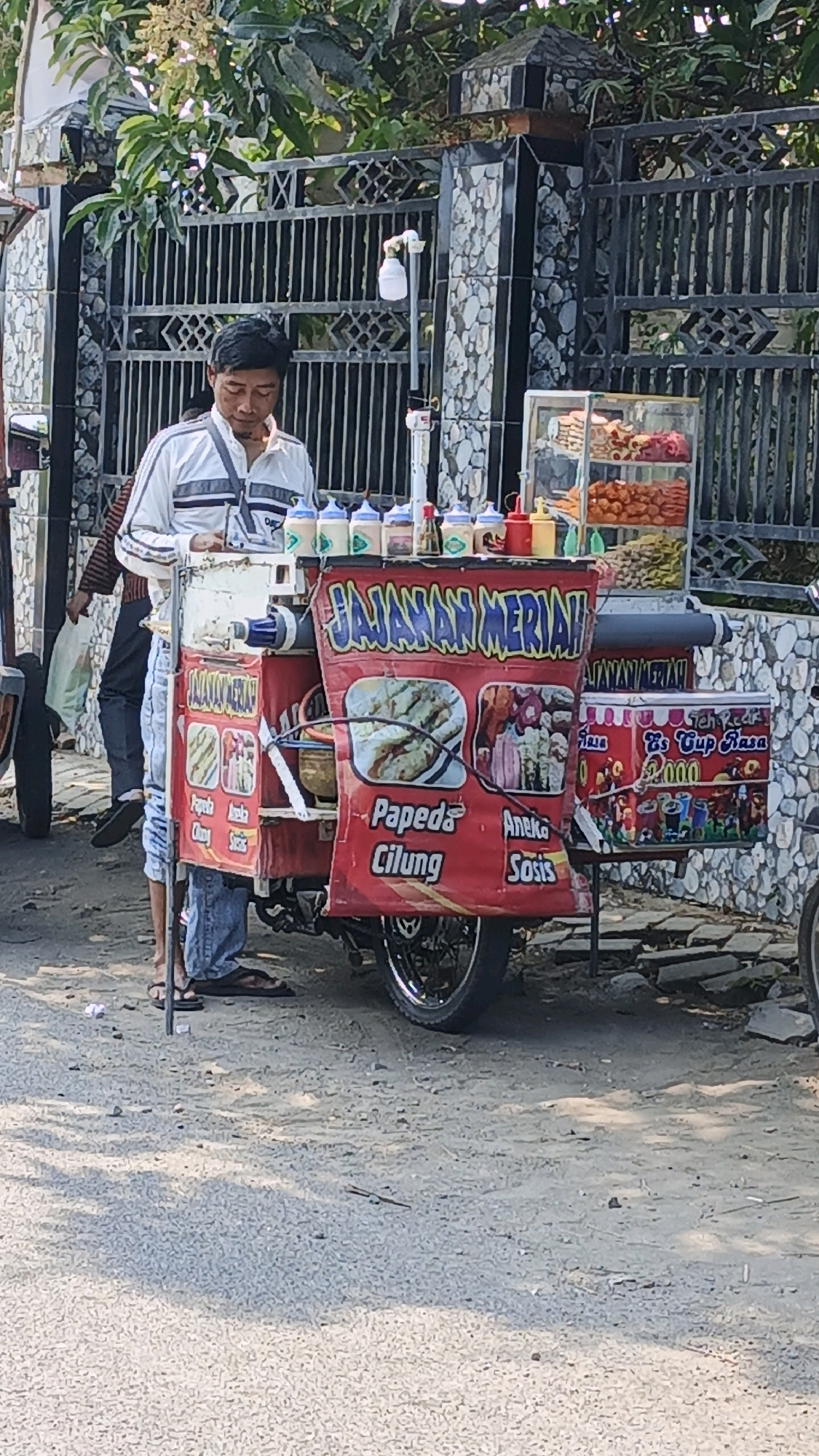 A man stands behind a food cart with a red tarp displaying various food items and prices. The cart is on a paved street in front of a decorative black metal gate and a stone-tiled wall. The man is wearing a white and gray striped long-sleeved shirt and blue jeans. He appears to be attending to the cart. The cart itself is equipped with bottles of condiments, what looks like ice cream cups for sale, and images of food items like "Papeda Cilung" and "Aneka Sosis." There are also colorful snacks displayed in a glass case on the cart. The overall scene suggests a street food vendor operating in an urban environment.