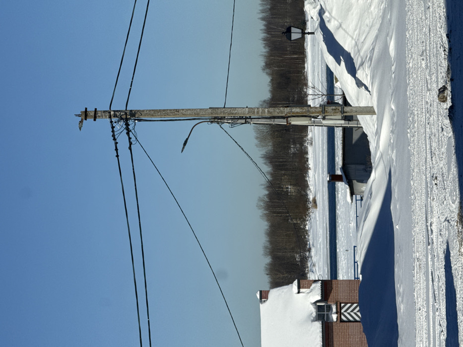 The scene depicts a winter landscape in Shlisselburg, Russia, under a clear, bright blue sky. The ground in the foreground is covered in white snow, displaying vehicle tracks and dark blue shadows cast by unseen objects to the right. To the left, a reddish-brown brick building with a pitched roof heavily covered in snow is partially visible. A window on this building features a vertical white and dark striped pattern directly beneath it.
Dominating the mid-ground is a tall, weathered utility pole, likely concrete or wood, from which numerous black electrical wires extend horizontally across the frame and diagonally downwards. A black street lamp fixture, curved downwards, is attached to this pole. Further to the right, a second, older-style street lamp with a dark, square-shaped lantern atop a pole is visible, surrounded by a large snowdrift.
Behind the utility pole, a smaller, low-slung building with a dark roof and a red brick chimney is partially obscured by snowdrifts. A wide body of water, partially frozen with patches of open, dark blue water and areas of white ice, stretches across the mid-background. Beyond the water, a dense line of bare, brown deciduous trees marks the distant bank, set against the low horizon. No individuals are present or engaged in any discernible actions.
