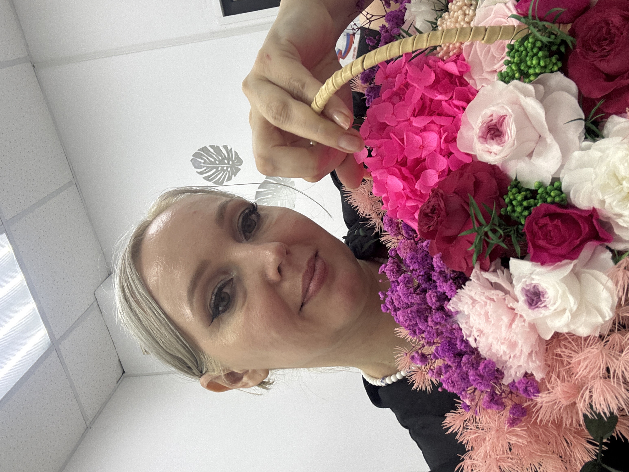 In Komsomolsk-on-Amur, Russia, a medium shot captures a woman with blonde hair, pulled back, and visible makeup, including eyeliner and mascara. She is wearing a black top and a white beaded necklace. Her right hand is holding the handle of a woven basket filled with a vibrant arrangement of flowers. The bouquet contains various blooms in shades of pink, purple, white, and magenta, including what appear to be hydrangeas, roses, and other smaller filler flowers. The background is a plain, white wall with a decorative grey leaf motif and a section of a white, tiled ceiling with fluorescent lighting. The overall scene suggests an indoor setting, possibly a home or a flower shop.