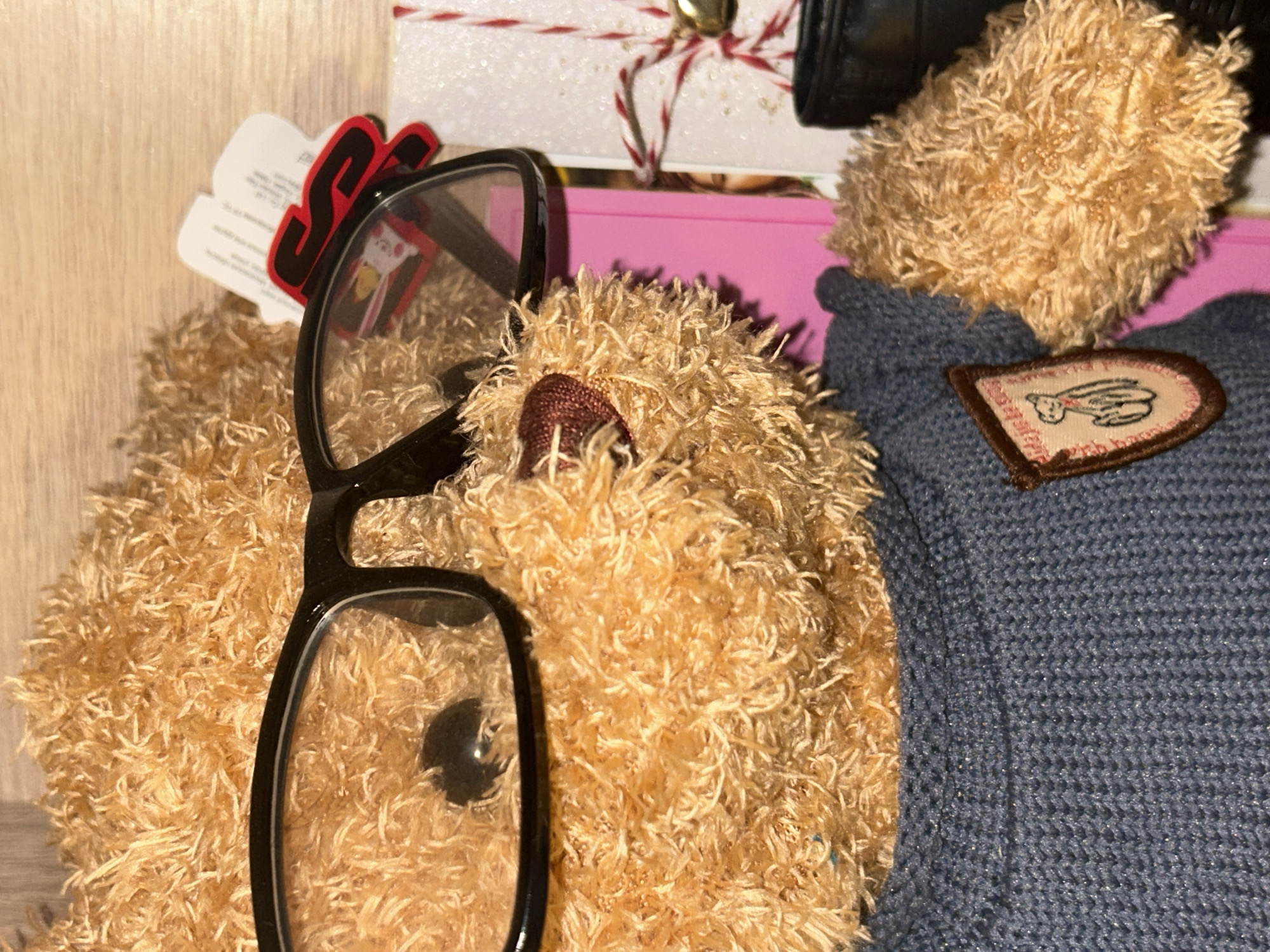 A close-up shot depicts a light brown teddy bear wearing black glasses. The teddy bear is also wearing a blue sweater with a brown patch featuring a graphic of a bear's head and paw. Behind the teddy bear, a white card with red and white twine, and a gold bell are visible. A pink rectangular object is also behind the teddy bear. The background consists of a light brown wooden surface. The location is unknown.