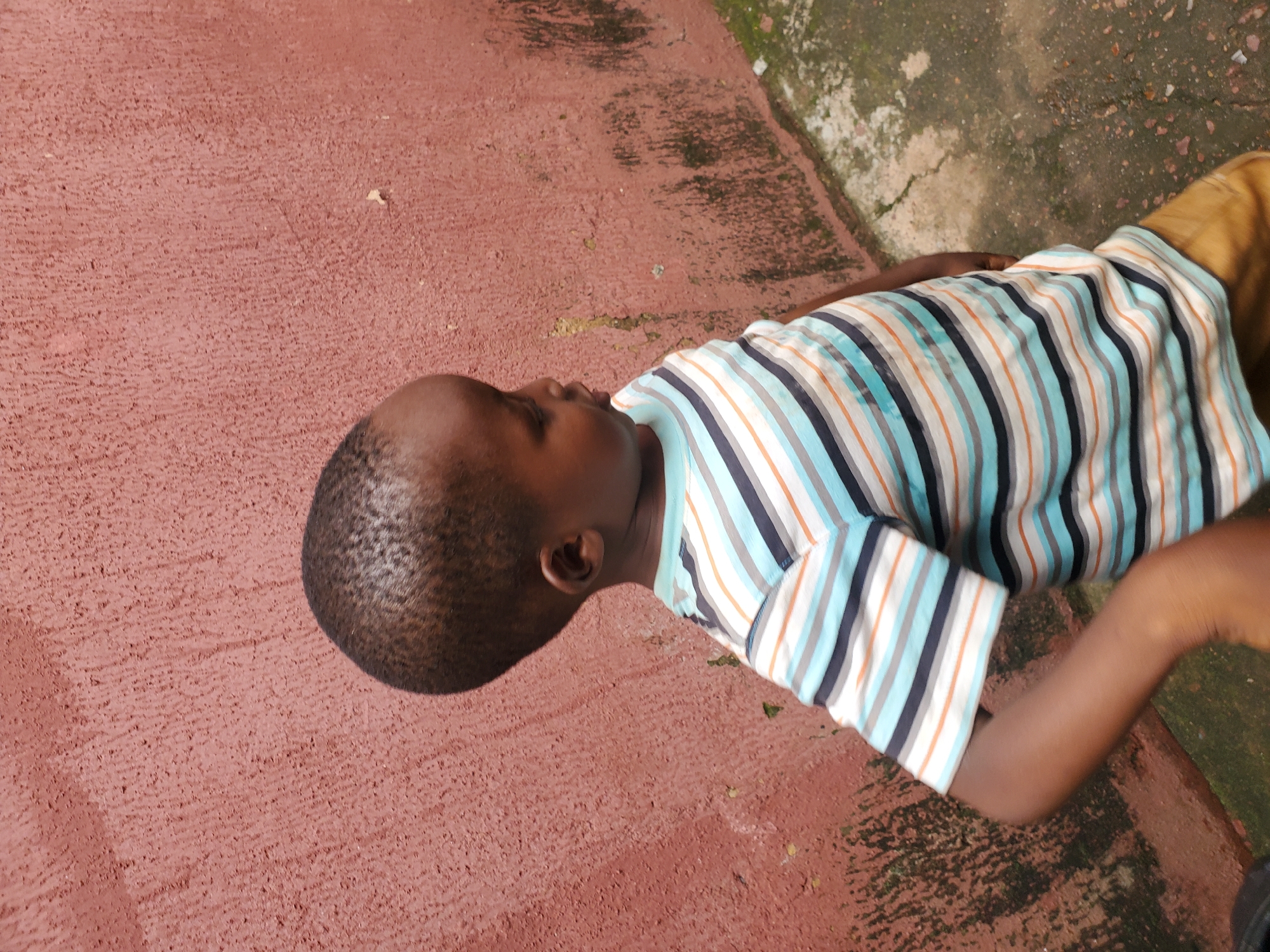 A child, viewed from above and slightly to the side, is lying on a reddish, textured surface. The child is wearing a horizontally striped shirt with alternating bands of white, blue, gray, and orange. The child's head is turned to the left, with their face and ear visible. Their skin tone is dark. The reddish surface appears to be concrete or asphalt and is mottled with some darker patches. To the right of the child, there is a darker, mossy or grimy area, suggesting an outdoor setting. The lighting appears to be natural, casting soft shadows. The child's expression is not clearly discernible. There is no visible text in the image.