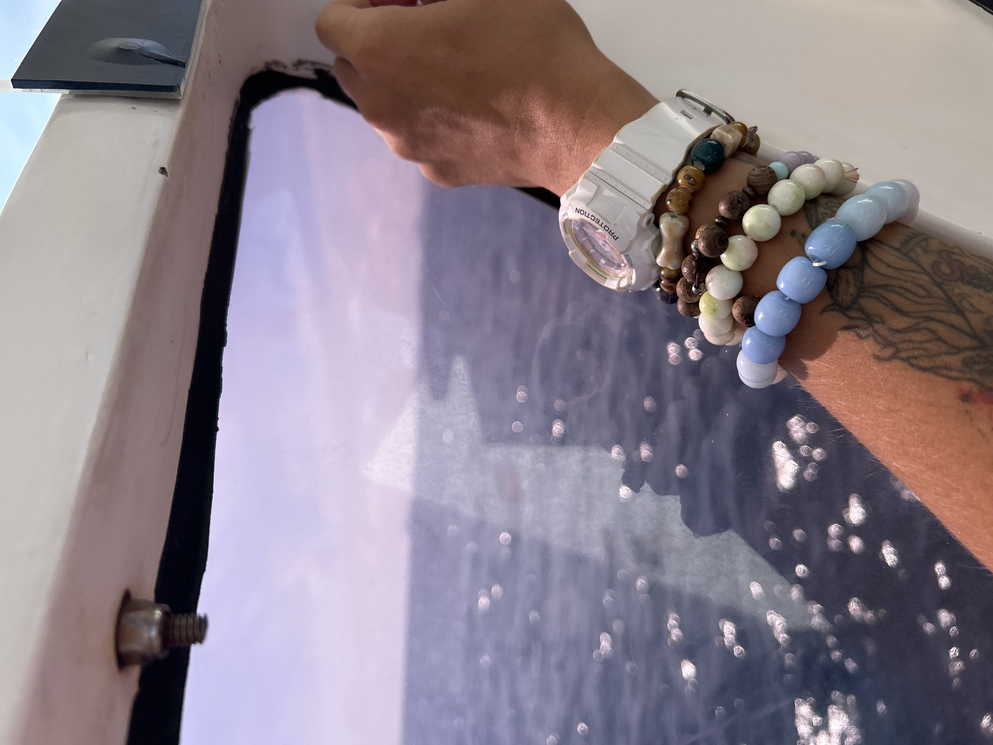 A close-up shot shows a person's left arm, adorned with multiple beaded bracelets and a white wristwatch with the word "PROTECTION" visible on its face, resting against a window. Outside the window, the ocean is visible, with sunlight glinting off the water's surface, suggesting a bright, clear day. The window frame is white, and a bolt protrudes from the top. The overall scene depicts a relaxed moment, likely on a boat or a waterfront establishment, offering a serene view of the sea. The photograph was taken in Rawai, Thailand, a location known for its coastal beauty and proximity to various islands and marine activities.