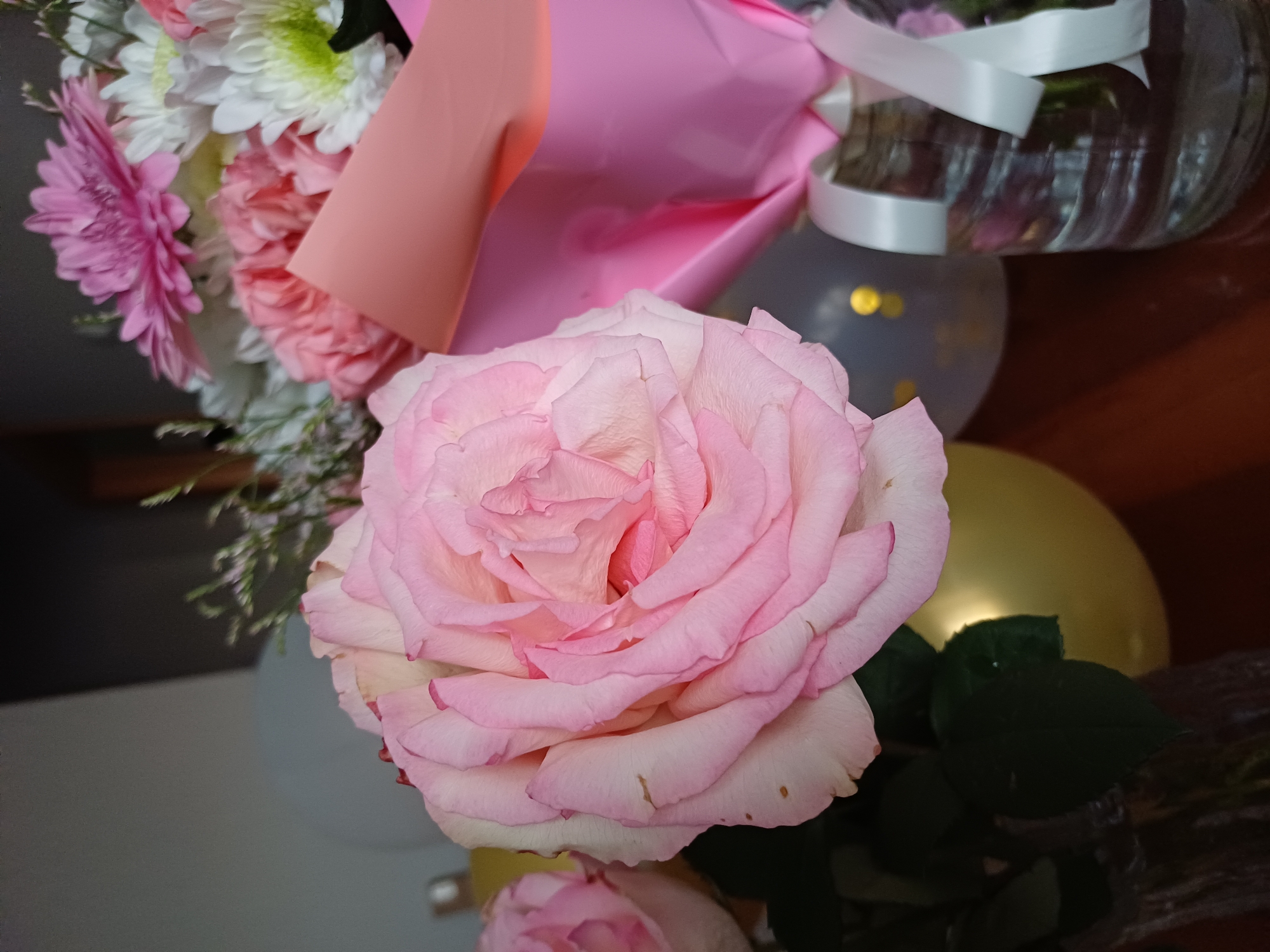 This image displays a close-up of a bouquet of flowers, with a prominent pink rose in the foreground. The rose is in full bloom, with layers of delicate pink petals. Behind the rose, other flowers are visible, including white and green chrysanthemums and pink carnations. The bouquet is partially wrapped in pink and peach-colored paper.
In the background, there are several balloons, one gray and one gold, suggesting a celebratory occasion. A glass vase with white ribbons tied around it is also visible. The overall setting appears to be indoors, possibly on a wooden surface, with a dark background. The lighting is soft, highlighting the textures and colors of the flowers. There is no visible text in the image. The image evokes a feeling of joy and festivity, likely related to a special event or gift.