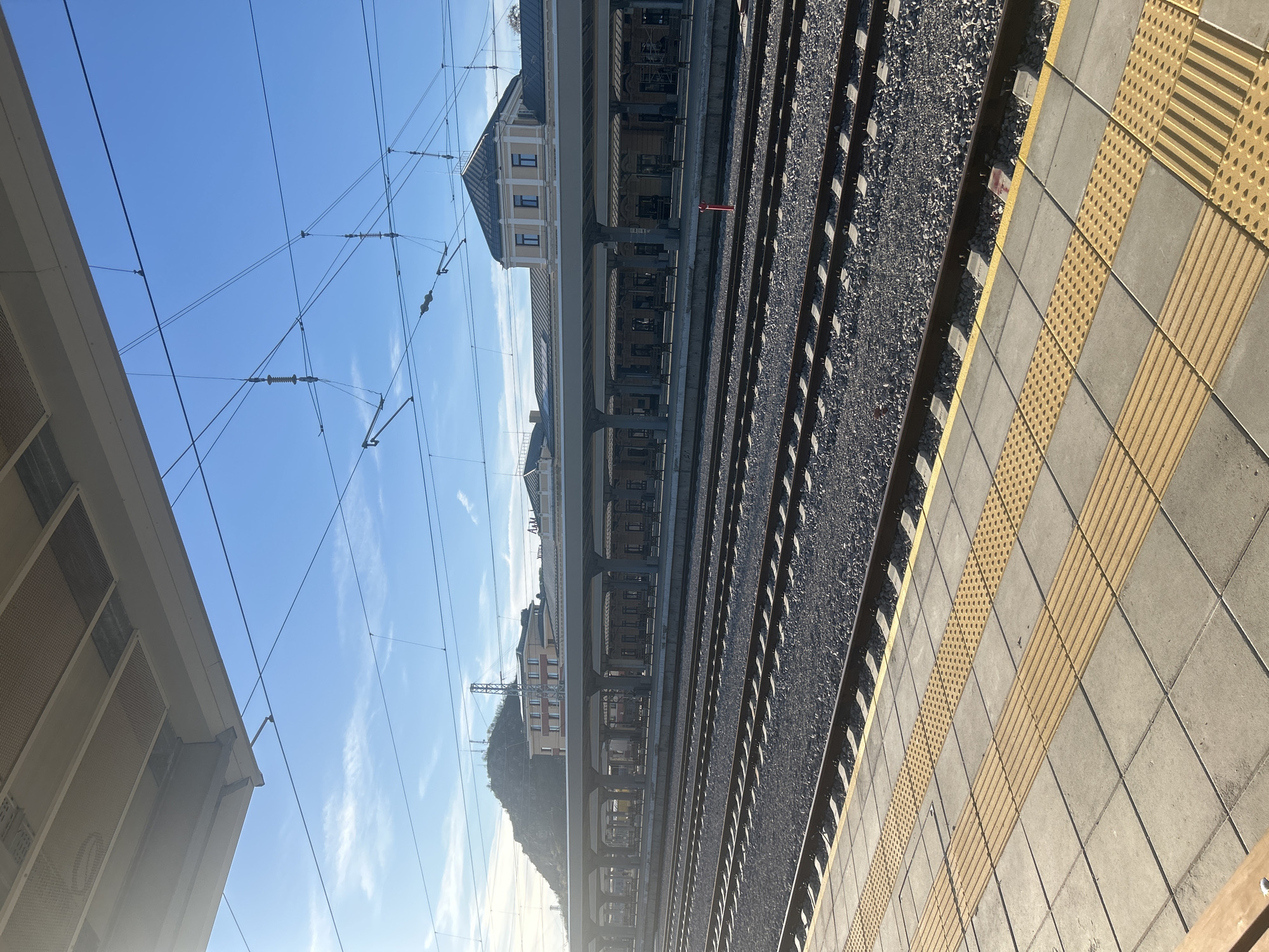 The image depicts a railway platform at the Plovdiv Central Railway Station in Bulgaria. The view is from platform level, looking across the train tracks towards the station building and beyond. The station building is multi-storied, with a light-colored facade and visible windows. Overhead power lines run along the tracks. The tracks themselves are surrounded by gravel. The platform edge is marked with tactile paving, consisting of patterned yellow and gray tiles. There are no visible people. The sky is clear and blue.