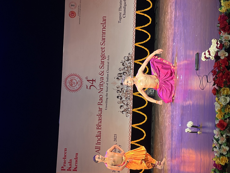 The image shows a performance at Tagore Theatre in Chandigarh, India. The stage is decorated with yellow garlands. Two male dancers are performing on stage. The dancer in the foreground is wearing a pink skirt and a golden belt. He is in a kneeling position, facing the audience. The dancer in the background is wearing a yellow skirt and a golden belt. He is standing, facing the audience. The stage is lit with purple and pink lights. There is a banner behind the dancers with the text "54th All India Bhaskar Rao Nritya & Sangeet Sammelan". There is also a sign in the bottom right corner of the image that says "Tagore Theatre Chandigarh". The coordinates (latitude: 30.733, longitude: 76.79) correspond to the location of Tagore Theatre in Chandigarh, India.