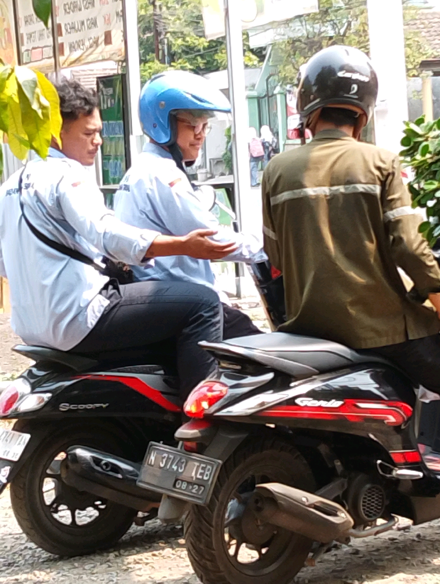Two men are on a black scooter with red and silver accents. The man in the front is wearing a light blue shirt and black pants, with a blue helmet. He is looking to his left. The man behind him is wearing a brown jacket and black pants, with a black helmet. He is looking straight ahead. The scooter has a license plate that reads "N 3743 TEB 08.27". The background shows a building with advertisements and some greenery. The scene appears to be in a city environment in Gempeng, Indonesia, likely a street or parking area.