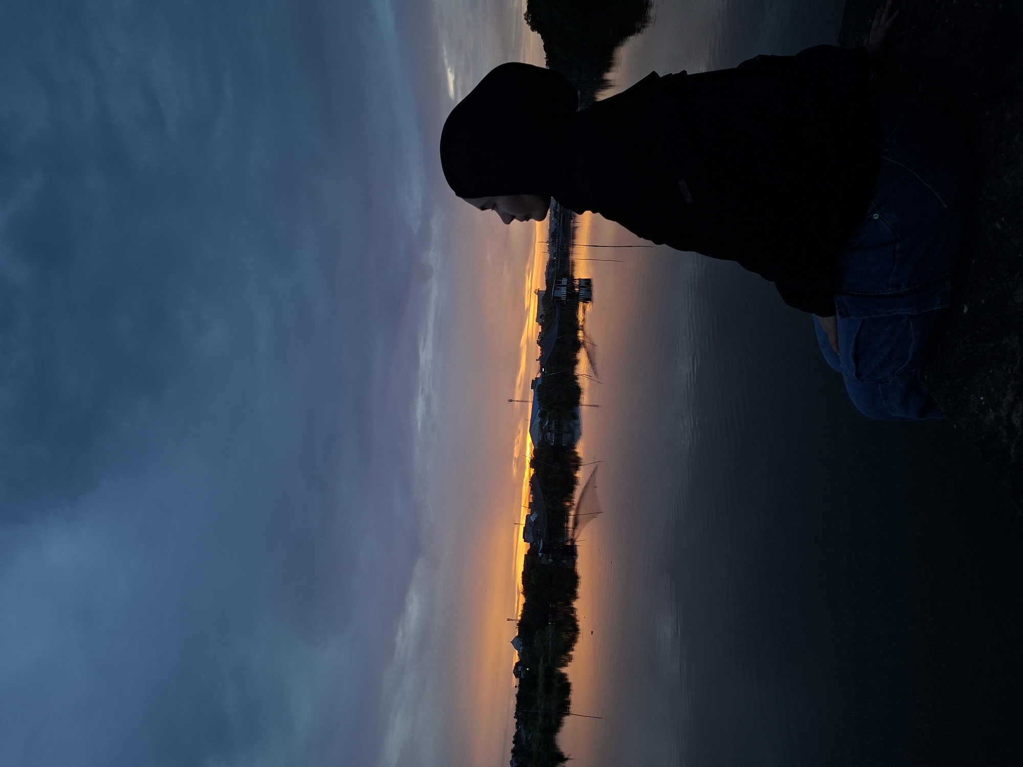 A person wearing a dark hooded jacket and blue jeans is crouched at the edge of a body of water at dusk. The sun is setting, casting an orange glow across the sky and water, which reflects the silhouette of buildings and trees. The sky transitions from a dark blue to orange near the horizon. The image appears to be taken in Lhokseumawe, Indonesia, based on the presence of buildings that match the architecture and characteristics of the city.