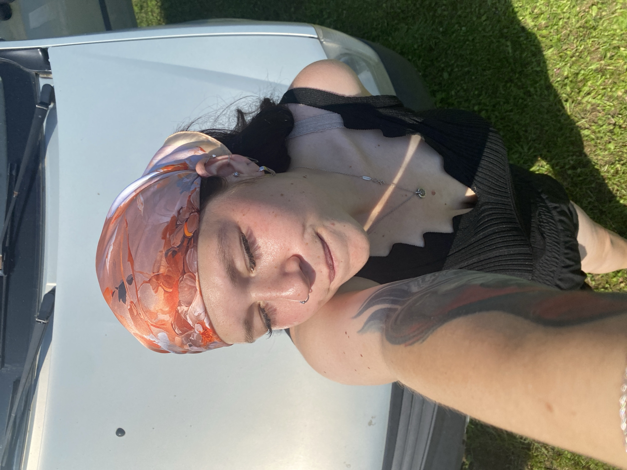 A young woman with dark hair, wearing a black ribbed tank top and an orange and white patterned headscarf, is photographed outdoors. She is wearing a nose ring and several earrings, including a hoop and studs. A silver necklace with a circular pendant rests on her collarbone. She has a tattoo on her left arm. The woman is leaning against the hood of a light-colored car, which is parked on a grassy area. The background suggests a sunny day. The image is an overhead shot, as if taken from a drone or by the subject herself looking up. The location is identified as Fokino, Russia.