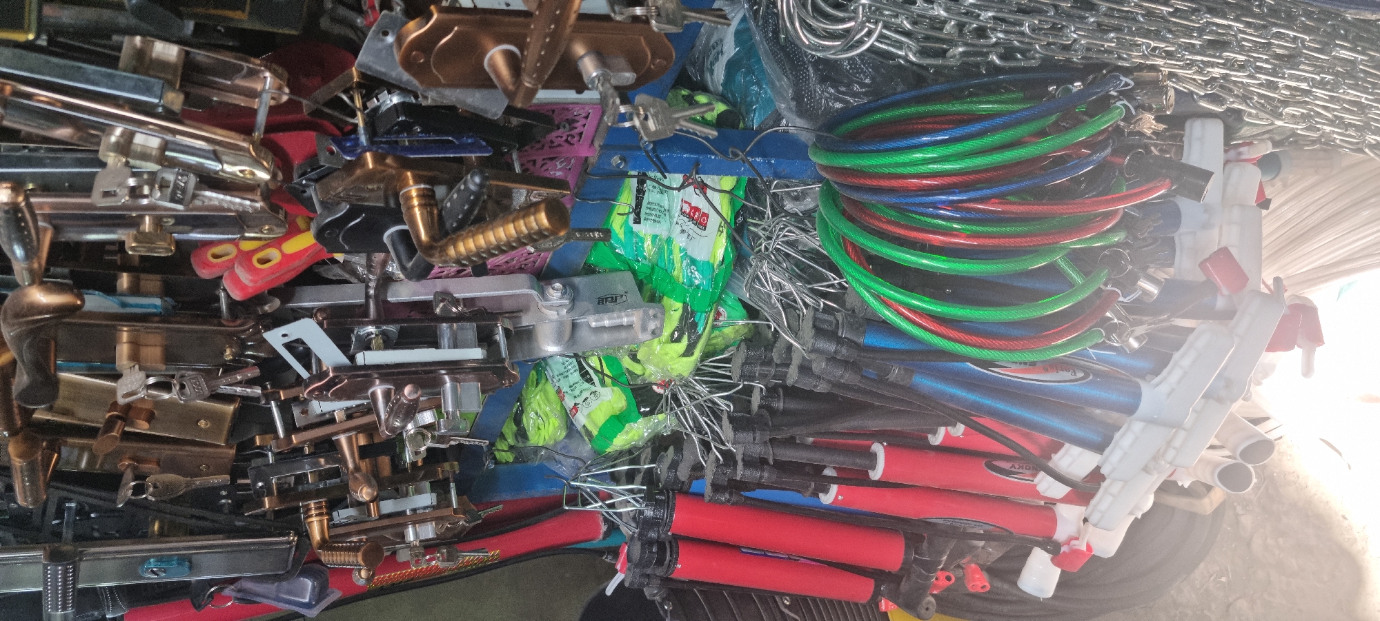 A close-up, high-angle, eye-level shot captures a dense display of hardware items for sale, likely in a market stall in Bokhtar, Tajikistan. The foreground is dominated by a jumble of various objects, including several red and blue cylindrical items, possibly bike pumps, with white plastic handles. Next to these, a collection of multi-colored coiled cables, predominantly red, green, and blue, are intertwined. These are likely security cables or locks, with several keys dangling from them. To the right of the cables, a heavy silver chain is visible.
The middle ground is filled with an assortment of door handles, locks, and keys. The handles are primarily made of brass and silver-toned metal, with some featuring decorative ribbed patterns. Several keys are attached to various locks, suggesting they are ready for purchase. Packs of bright neon green items, possibly gloves or other protective gear, are also visible, partially obscured by the hardware.
The background is a dense arrangement of more hardware, primarily door locks and handles, with a repeating pattern of pink and metallic elements that might be decorative inserts or part of the display structure. The overall impression is of a well-stocked, somewhat disorganized retail space specializing in security and building hardware. The lighting suggests an indoor setting, possibly with artificial light sources, casting some glare and shadows across the scene. No people are visible in the image.