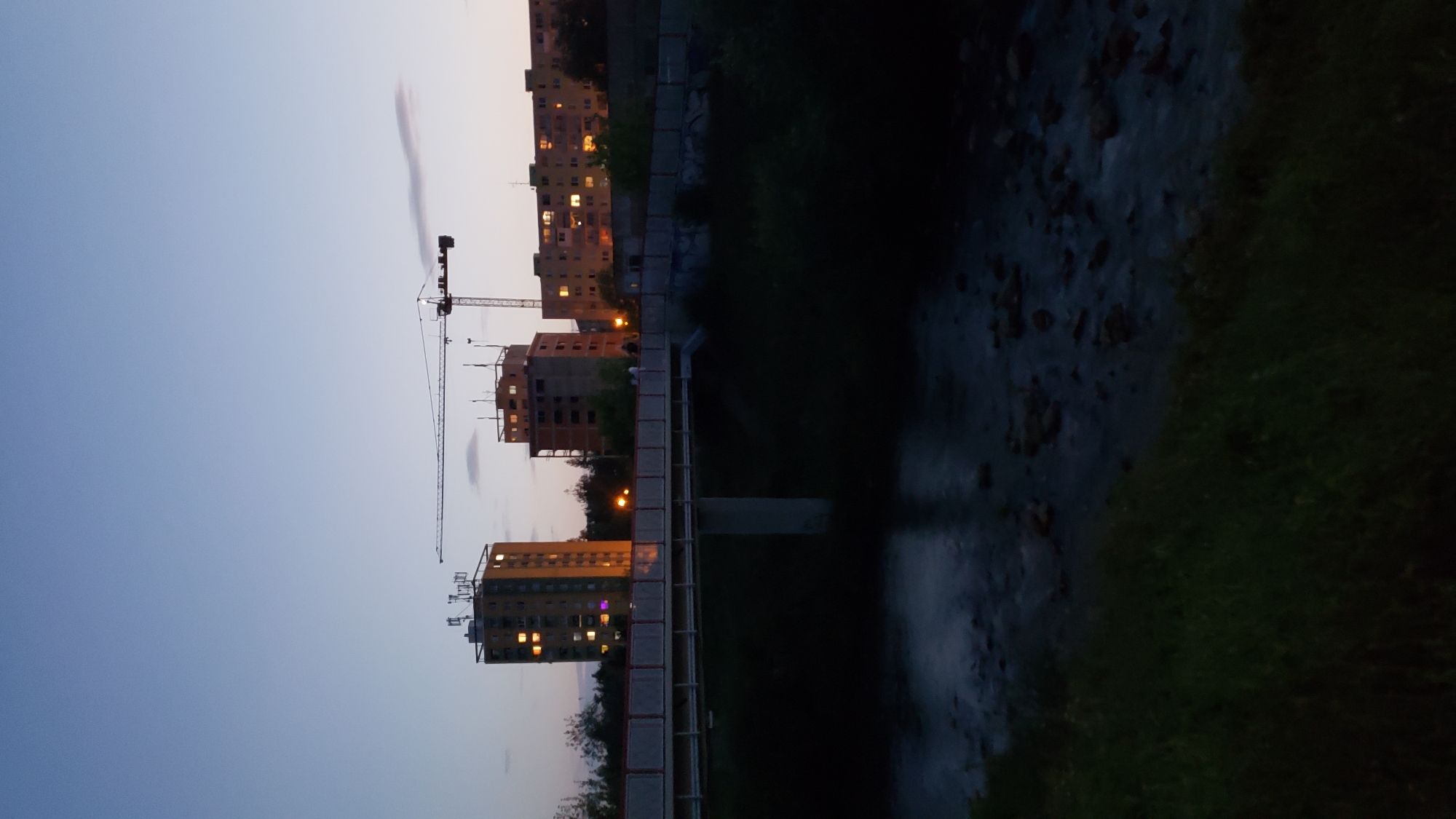 The image depicts a scene at dusk in Prešov, Slovakia, near the Sekčov district. In the background, several multi-story residential buildings are visible, with lights illuminated in some of the windows, suggesting occupancy. A prominent construction crane stands to the right of the central buildings, indicating ongoing development or renovation. In the foreground, a bridge spans across a body of water, likely a river or stream, with visible rocks and ripples on the surface. The banks of the water body are covered in dark vegetation, transitioning to grass in the lower part of the frame. A few indistinct figures are present on the bridge, suggesting pedestrian activity. The overall atmosphere is subdued due to the low light conditions.