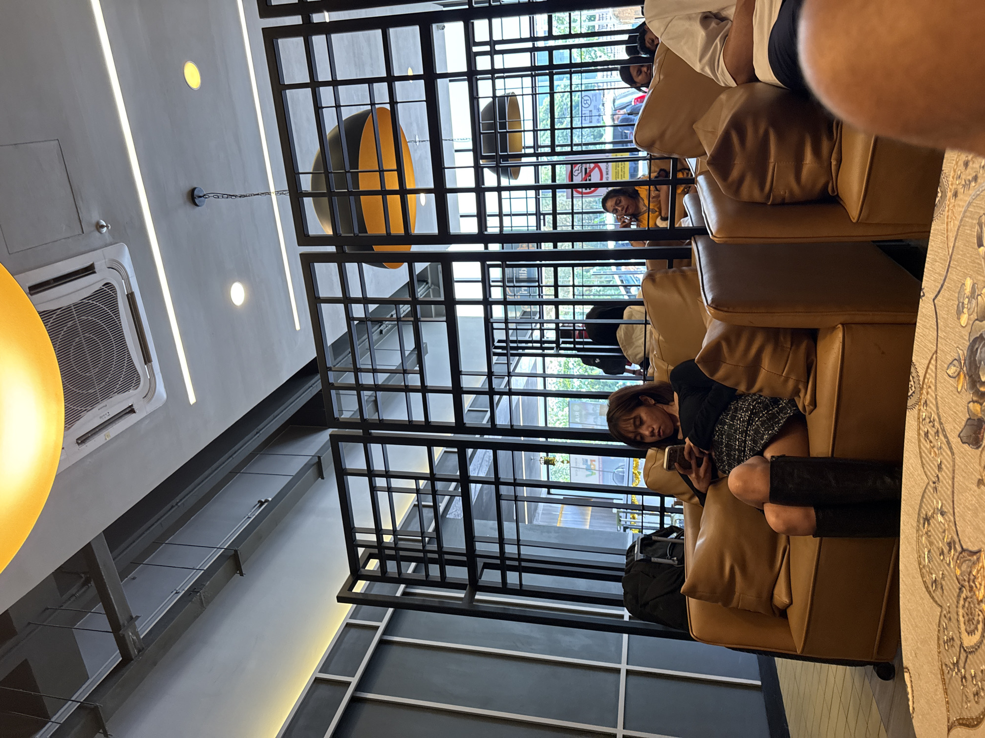 The image depicts an interior space, likely a café or lounge, in Kuala Lumpur, Malaysia. A black metal grid structure partially obscures the view of the outside. A woman is reclined on a brown leather sofa, looking at a smartphone. A black backpack sits beside her. Several other individuals are present in the background, some on their phones and having conversations. Lighting fixtures are visible on the ceiling.