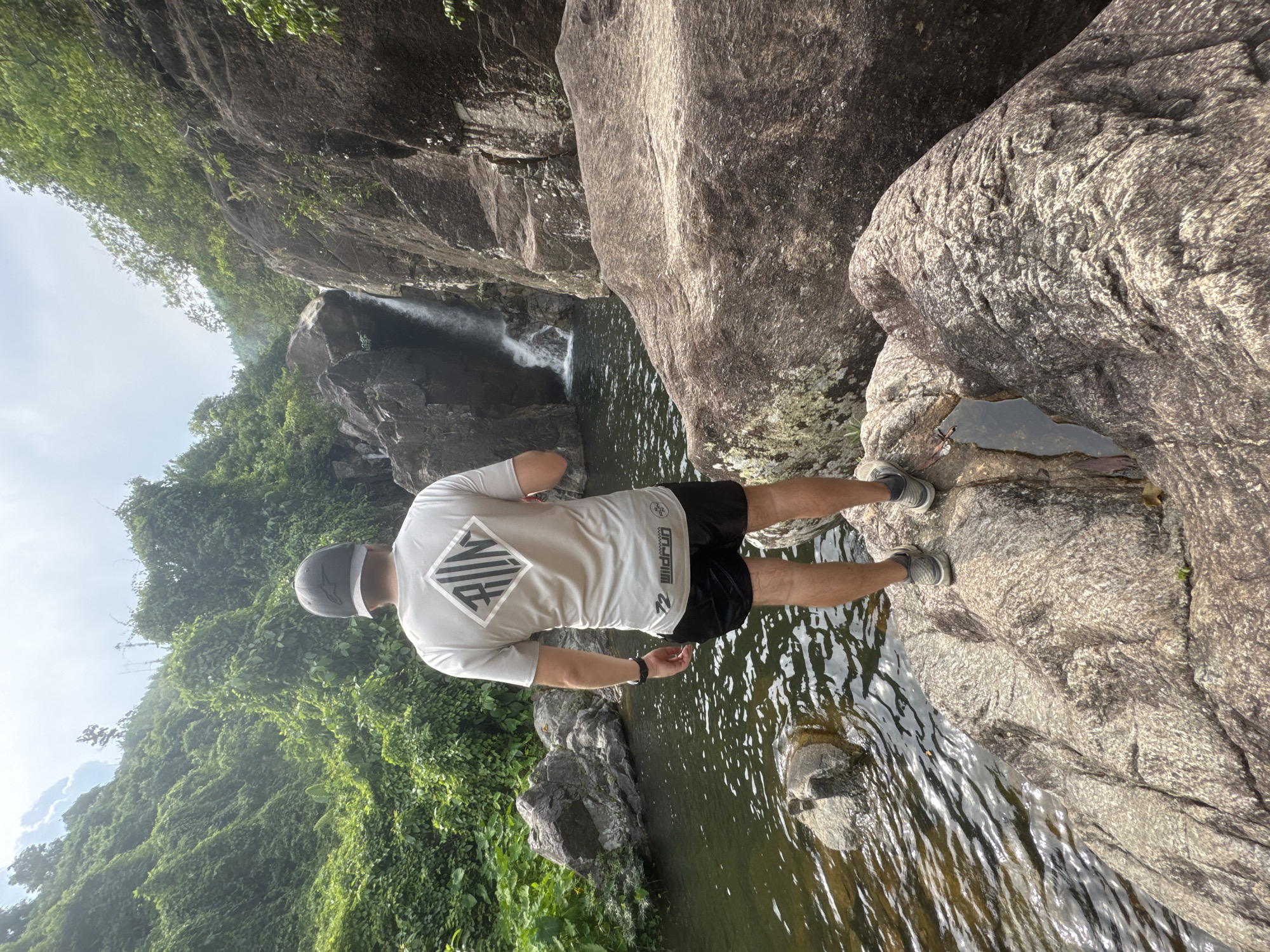 The image shows a person standing on rocks near a waterfall in what appears to be a mountainous or forested area. The person is wearing a light-colored shirt with a graphic on the back, dark shorts, a hat, and athletic shoes. They are standing near the edge of a stream, with water flowing around the rocks. In the background, a small waterfall flows into a pool of water. Lush green vegetation covers the surrounding slopes. Based on the context clues, the location is likely in the vicinity of Đà Nẵng, Vietnam, which is known for its mountainous terrain and natural beauty.