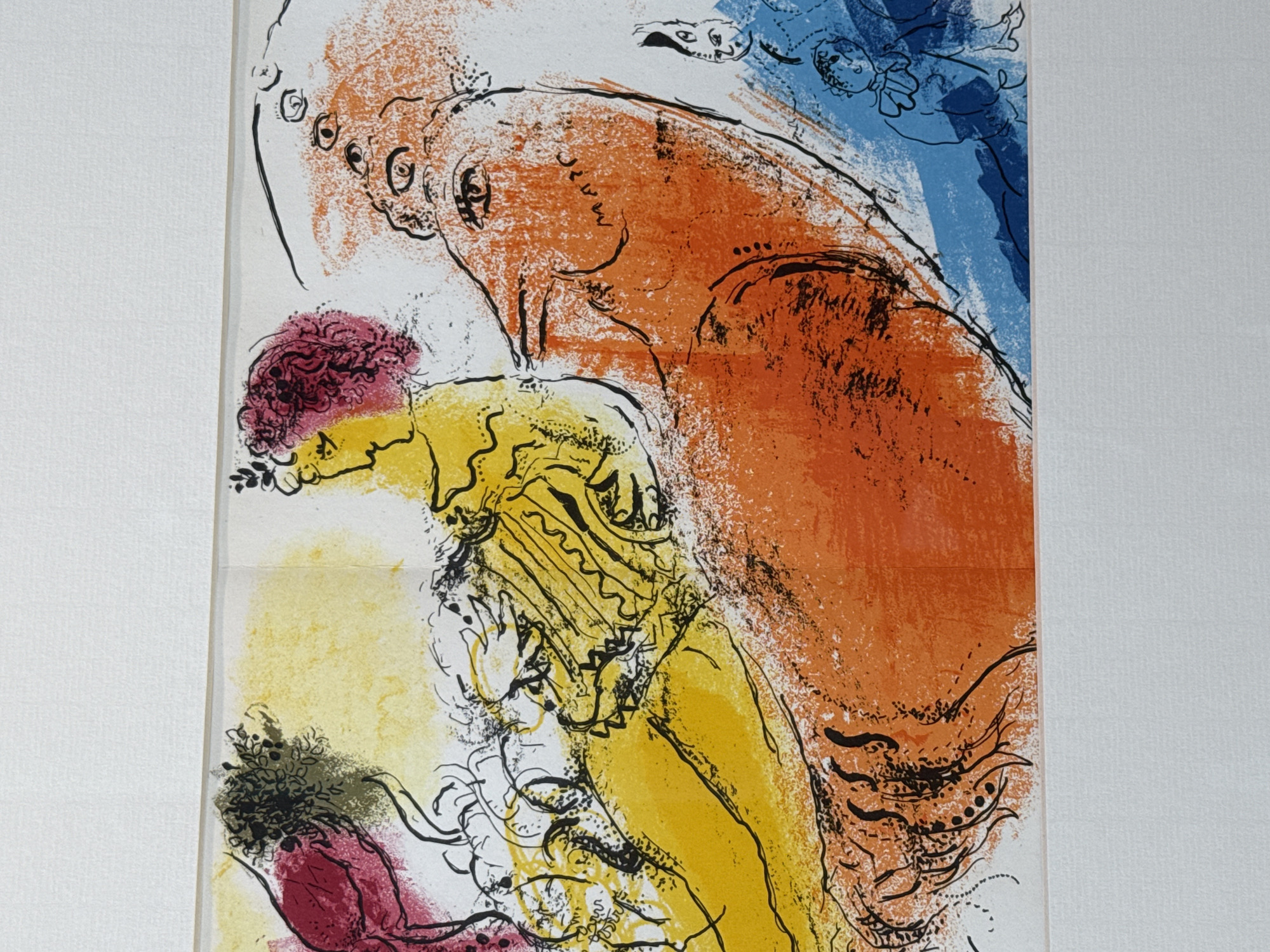 This is a close-up, eye-level, medium shot of a colorful lithograph by Marc Chagall, framed and displayed on a white wall. The artwork features a surreal scene with a woman with pink hair in profile, sitting amidst abstract, bold shapes and lines. A large, orange, creature-like form dominates the right side of the composition, with multiple eyes depicted. The creature's head is turned to the left, facing the woman. There are smaller, stylized figures in blue and black on the far right and bottom right. The overall style is expressive and dreamlike, with vibrant yellow, orange, pink, and blue colors. The lithograph appears to be a limited edition print, signed by the artist with "MARC" visible in the central orange area. The background of the artwork is white, and the framing suggests it is a gallery or home setting.