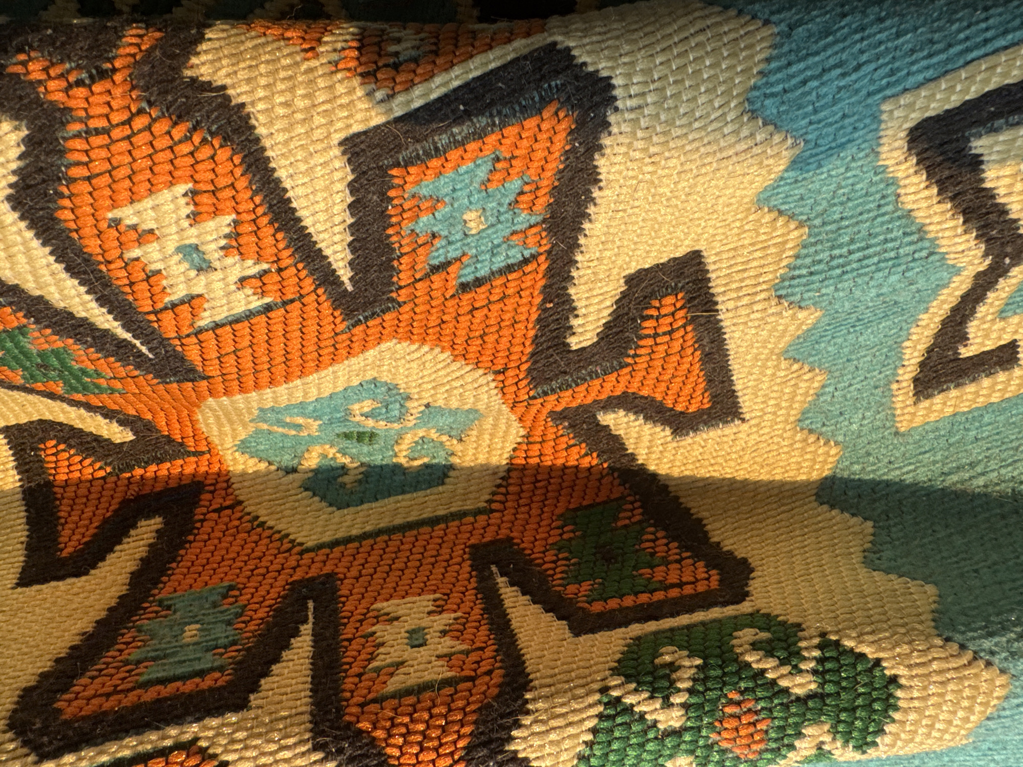 The image shows a close-up view of a woven textile, likely a rug or tapestry, with a geometric pattern. The dominant colors are orange, cream, black, teal, and green. The texture of the weave is clearly visible, with small loops and raised threads creating a tactile surface. The pattern features star-like motifs and zig-zag lines. The lighting suggests natural sunlight casting shadows, creating a sense of depth and highlighting the fabric's texture. The textile appears to be folded or draped, with some areas in shadow and others brightly illuminated. There are no people, animals, or any identifiable elements of a specific location within the city of Las Rozas de Madrid, Spain. The image is solely focused on the intricate details and patterns of the textile itself.