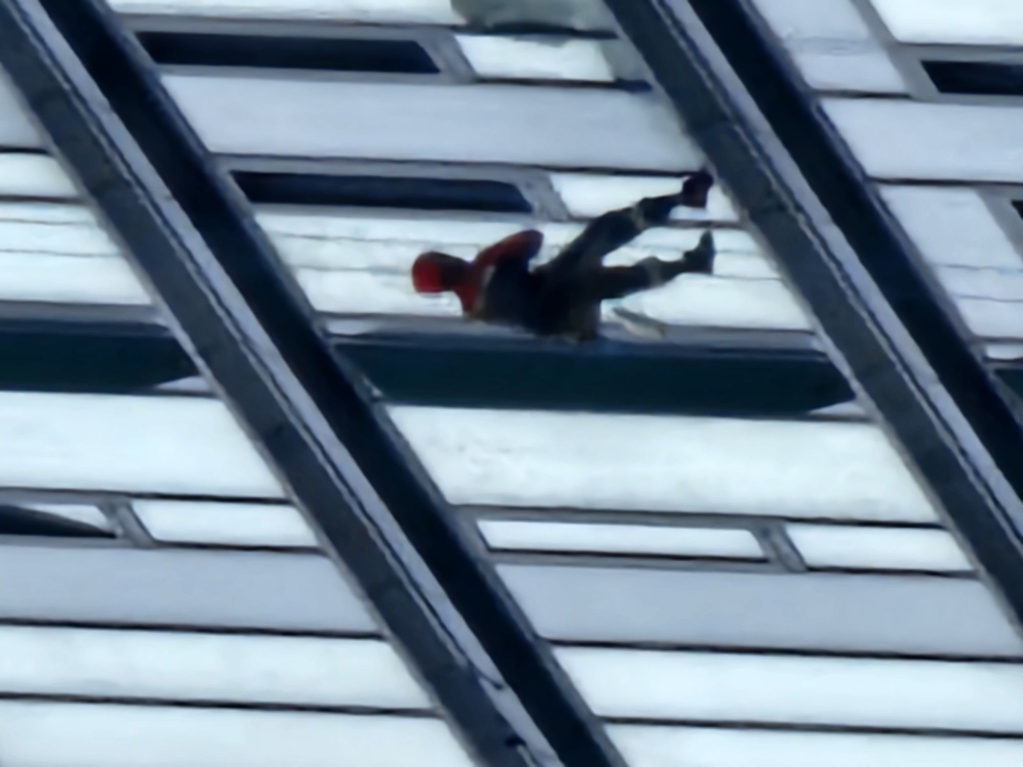 A person in a red and dark colored suit, resembling a superhero costume, is scaling the side of a modern building. The individual is positioned on a horizontal ledge, with their body angled outwards. The building's facade is composed of vertical panels, alternating between light and dark shades, and horizontal dark-colored bands. The person's limbs are bent as if in motion. The background shows more of the building's structure, with repeating patterns of windows or panels. The image appears to be taken from a distance, suggesting a city environment. The specific location in Moscow, Russia, is not identifiable from the visual information presented in the image.