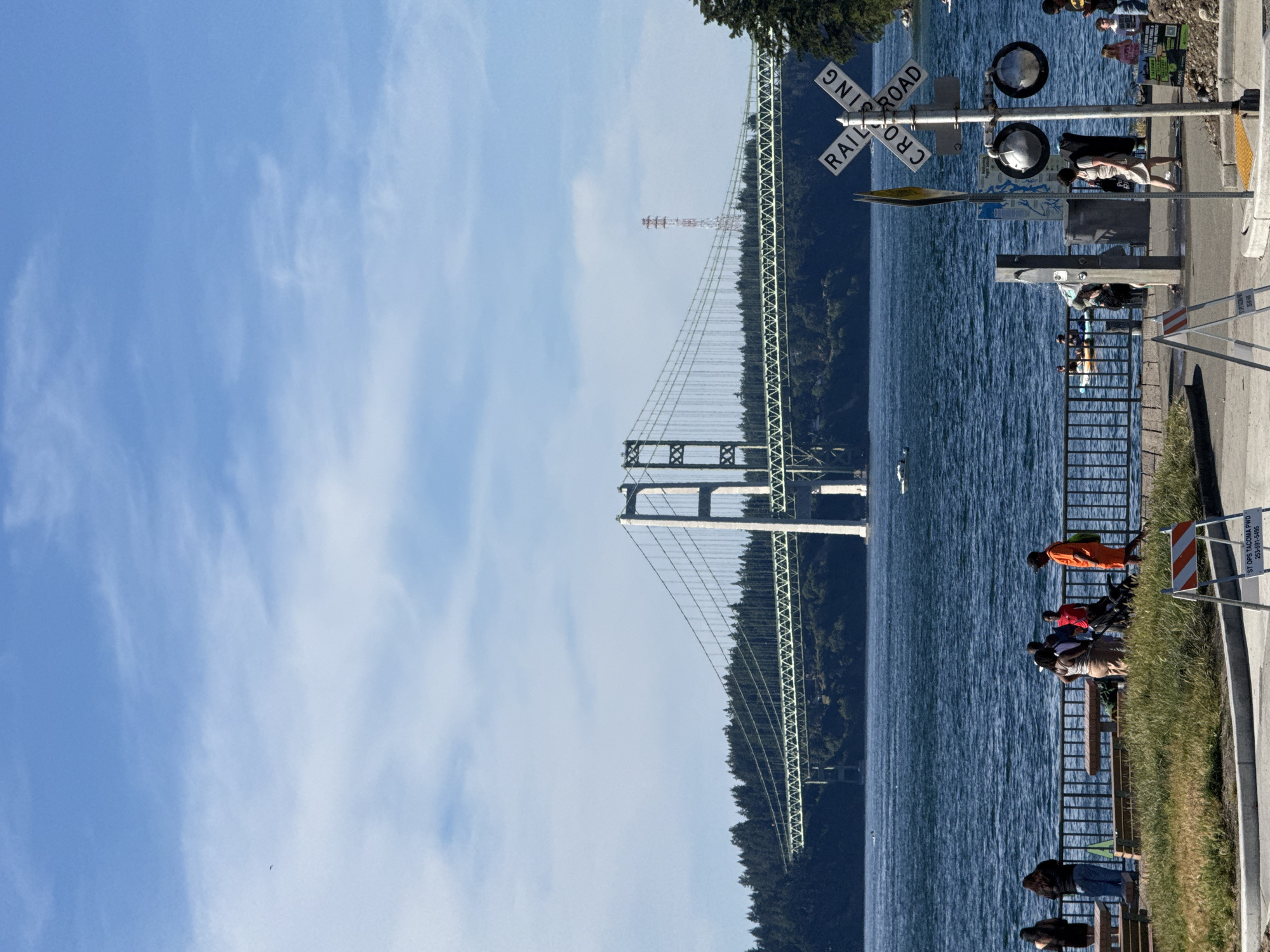 In Tacoma, Washington, a panoramic view captures the iconic Tacoma Narrows Bridge, spanning across the shimmering blue waters of Puget Sound under a bright, partially cloudy sky. The majestic suspension bridge, with its distinctive green towers and cables, is a prominent feature, connecting the mainland to the Kitsap Peninsula.
Along the waterfront, a diverse group of people are enjoying a sunny day. Some are gathered near picnic tables, while others stroll along the boardwalk, observing the serene water. A few individuals are relaxing on benches, taking in the scenic vista. The presence of a railroad crossing, complete with warning lights and signs, indicates the proximity of a railway line. The surrounding area is dotted with lush green grass and trees, adding to the natural beauty of the scene.