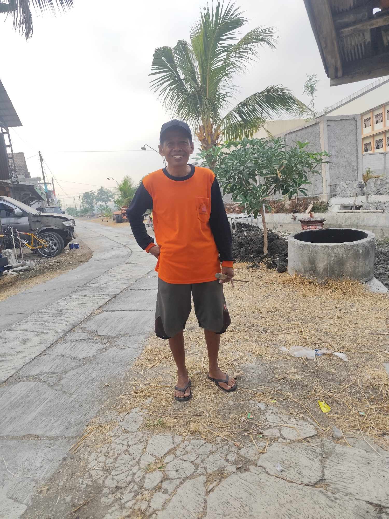 A man stands on a cracked concrete pathway in a semi-rural area, likely Purwodadi, Indonesia. He is wearing an orange long-sleeved shirt with black sleeves and dark gray shorts, along with black flip-flops. He has a black baseball cap on and is smiling at the camera, holding a small tool in his right hand. The environment appears to be an outdoor construction or renovation site. To the man's left, a section of the pathway is bordered by a ditch. Behind him, a palm tree, a small leafy tree, a concrete well, and piles of dirt are visible. In the background, a damaged car and a bicycle are parked along the pathway. Further in the distance, a building with multiple windows and a grey concrete wall can be seen. The sky is overcast.