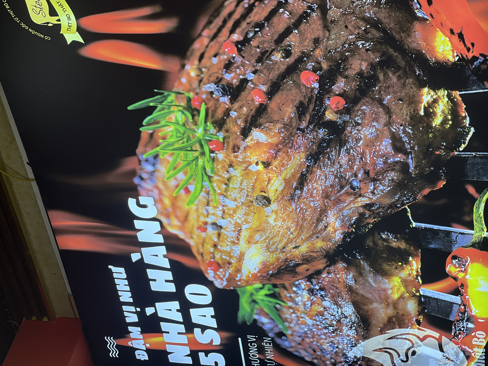 The image shows a promotional poster, likely for a steak restaurant in Đà Nẵng, Vietnam. The poster features a close-up of a grilled steak, garnished with rosemary, peppercorns, and salt. The steak is positioned on a grill with visible flames in the background. Vietnamese text on the poster reads "ĐẬM VỊ NHƯ NHÀ HÀNG 5 SAO" (translating to "Rich taste like a 5-star restaurant") and "HƯƠNG VỊ TỰ NHIÊN" ("Natural Flavor"). A ribbon on the top left says "THỊT BÒ THẬT" ("Real Beef"). The steak appears juicy and well-seasoned, visually emphasizing the restaurant's quality.