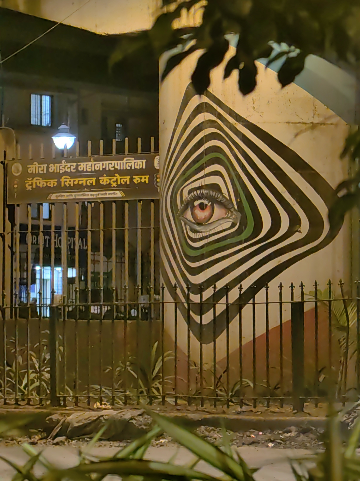 The image shows a mural of an eye within a spiral pattern painted on a concrete pillar, located in Mira-Bhayander, India. The pillar is part of a structure that appears to be a building entrance or a boundary wall. Behind the pillar, a dark metal fence encloses an area with some foliage. A sign above the fence, written in Marathi, indicates "Mira-Bhayander Municipal Corporation: Traffic Signal Control Room." Below this sign, the text "पुरविल्या आणि अद्ययावत वाहतूक नियंत्रणासाठी समर्पित" is visible. Through the fence, a building with windows and a street lamp are discernible. The foreground shows the ground with scattered debris and some green plants. The scene is illuminated by artificial light, suggesting it is nighttime or a dimly lit area.