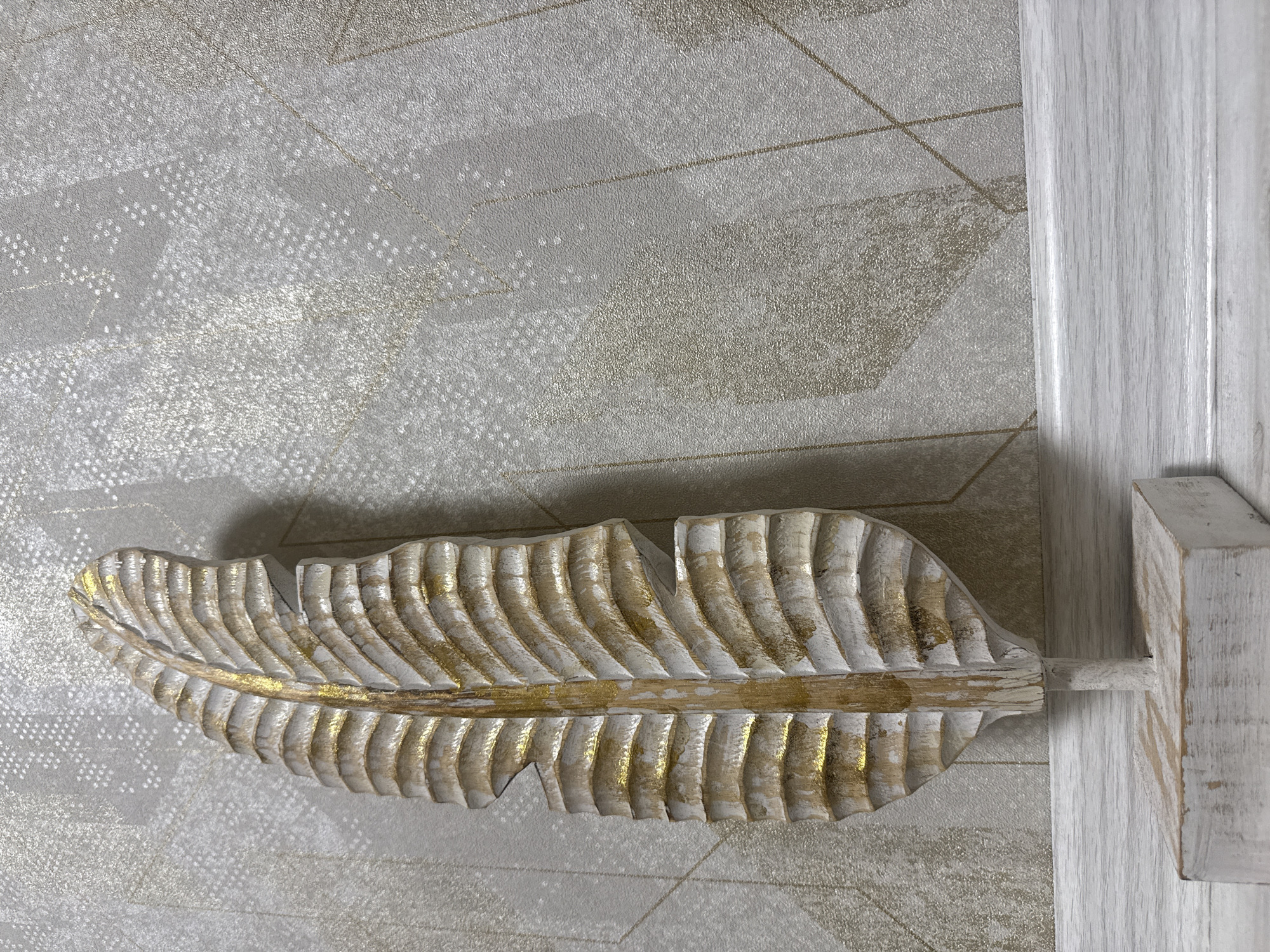 A close-up shot displays a decorative wooden feather mounted on a wall with patterned wallpaper, likely indoors. The feather is painted white with gold highlights, and it is attached to a white wooden fixture that is fixed to a white wooden panel on the right edge of the frame. The wallpaper features a geometric pattern in shades of gray and gold.